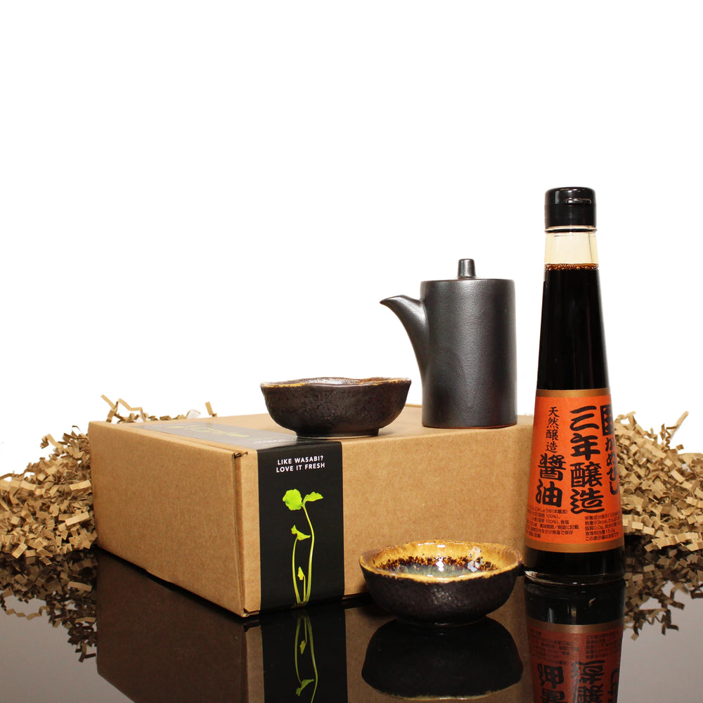 The 3 Year Aged Soy Sauce with Pourer & Dishes set, featuring a bottle of soy sauce, black ceramic pourer, two small dishes, and a cardboard box with brown paper filler, is displayed against a white background.