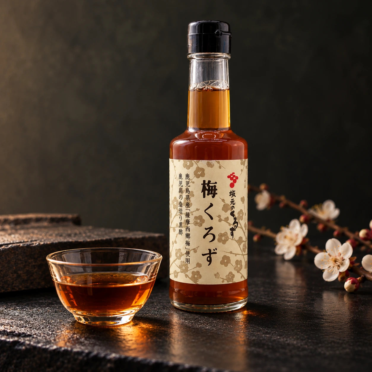 
                  
                    A glass bottle of Ume Kurozu (black rice vinegar with ume plum) - 200 ml and a matching cup filled with the amber-colored vinegar, set on a dark textured surface next to a plum blossom branch.
                  
                