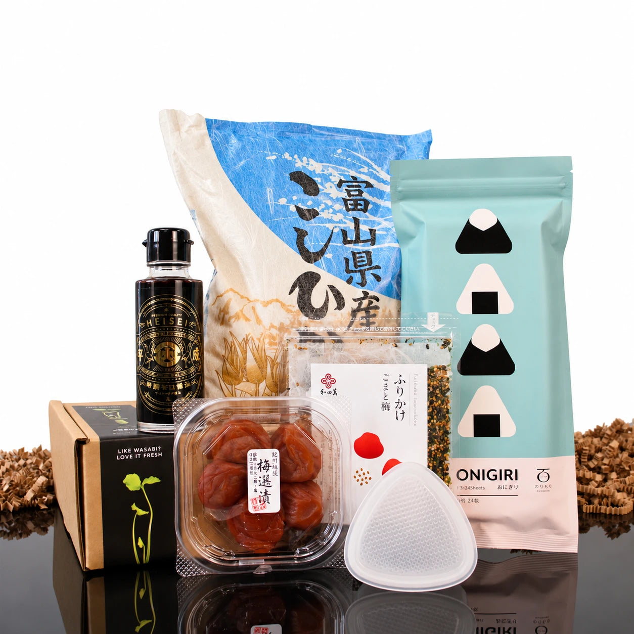 A Japanese Onigiri Kit including a bag of rice, nori sheets, soy sauce, pickled plums, seasoning, and a triangular mold for making rice balls.