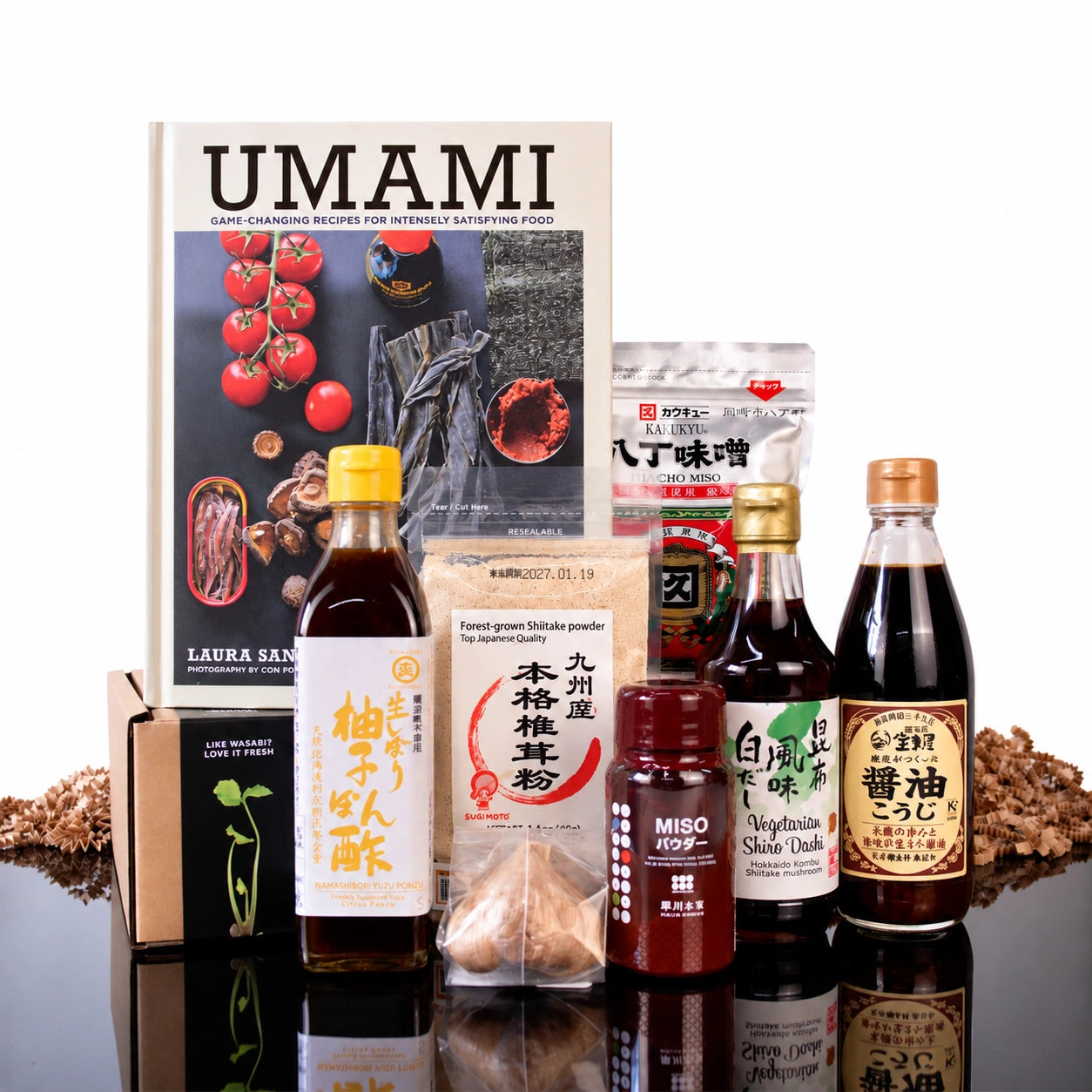 The Japanese Umami Box With Umami Cook Book displayed behind a Japanese pantry set of yuzu ponzu, dashi, soy sauce, miso, and shiitake powder.