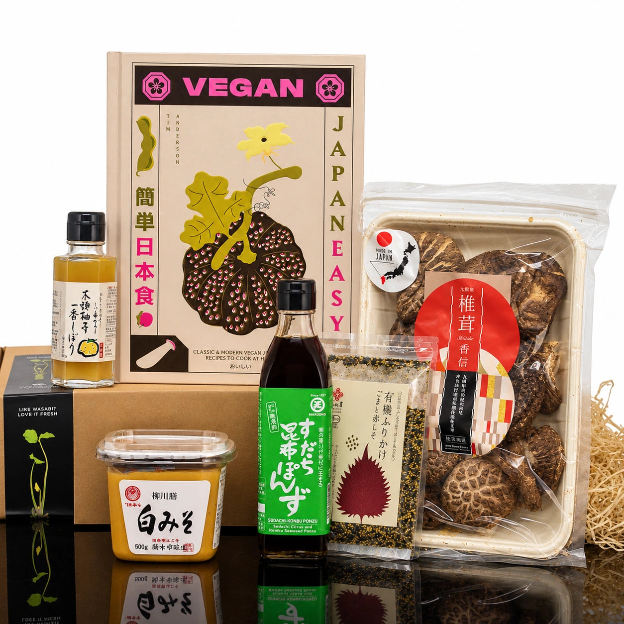 A Vegan Japanese Ingredients Set with Vegan Cook Book surrounded by yuzu juice, white miso, sudachi ponzu, dried shiitake mushrooms, and a seasoning packet.