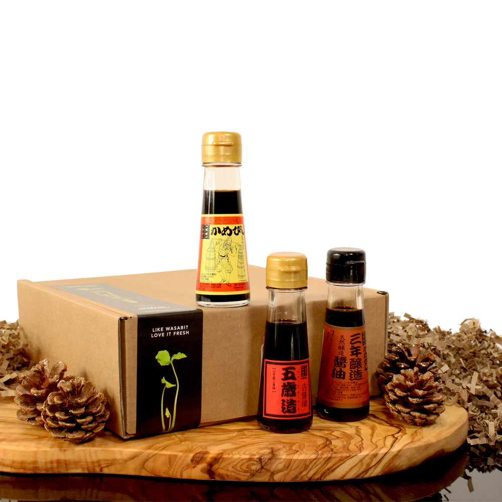 The Aged Soy Sauce Triple Pack is displayed on a wooden board with pinecones beside a brown box, set against a white background, showcasing the rich umami flavor of these three premium bottles.