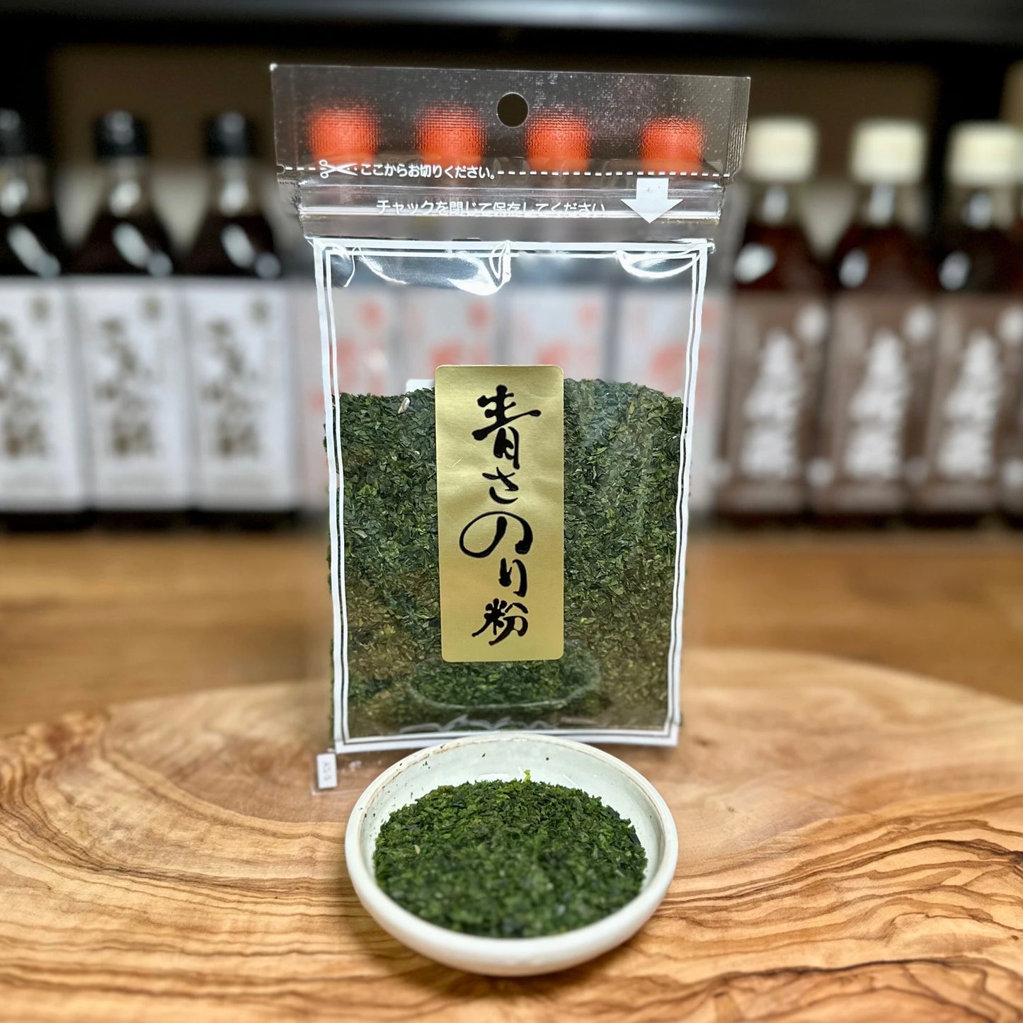 
                  
                    A clear plastic bag of Dried Aonori Seaweed Flakes - 20g stands on a wooden surface, accompanied by a small white dish of the seaweed; bottles with Japanese labels are visible blurred in the background.
                  
                