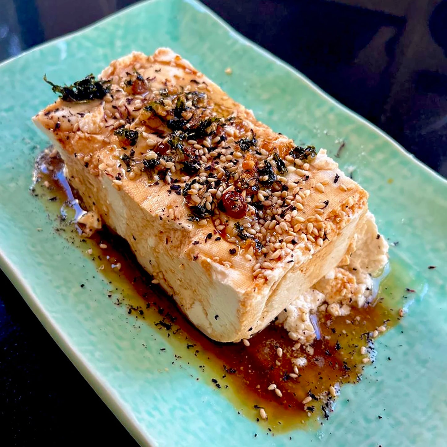 
                  
                    A block of tofu on a green plate, topped with sesame seeds, dried herbs, Aosa Seaweed Crispy Condiment - 90g, and a soy-based sauce.
                  
                