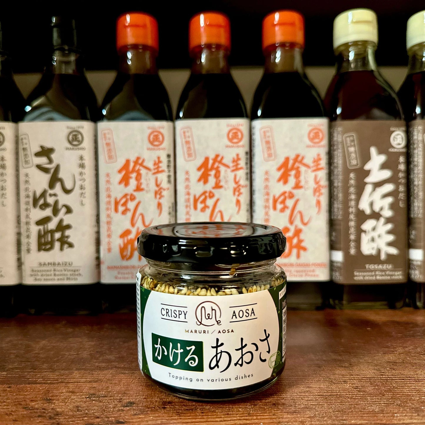 
                  
                    A jar of Aosa Seaweed Crispy Condiment - 90g, ideal as a crunchy topping, sits on a shelf in front of bottles of Japanese condiments such as Sanbanzu, Tosazu, and ponzu.
                  
                