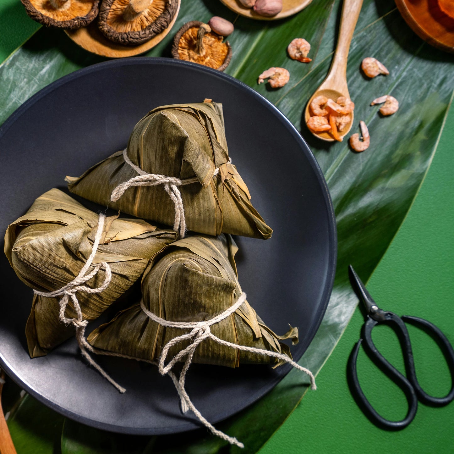 
                  
                    Three leaf-wrapped rice dumplings on a black plate, surrounded by dried ingredients, capture the tropical allure of the Japanese Hardy Banana Plant (Musa basjoo), evoking a vibrant garden setting.
                  
                