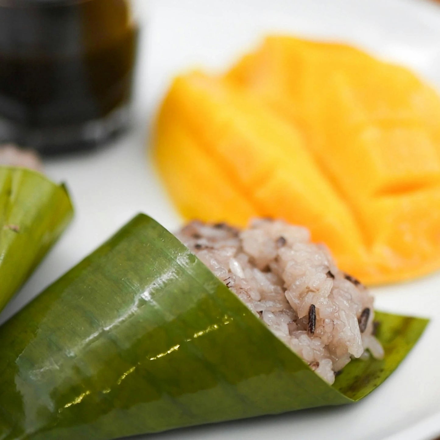 
                  
                    A close-up of sticky rice wrapped in a banana leaf from the Japanese Hardy Banana Plant (Musa basjoo), sliced mango, and a glass of dark liquid on a white plate.
                  
                