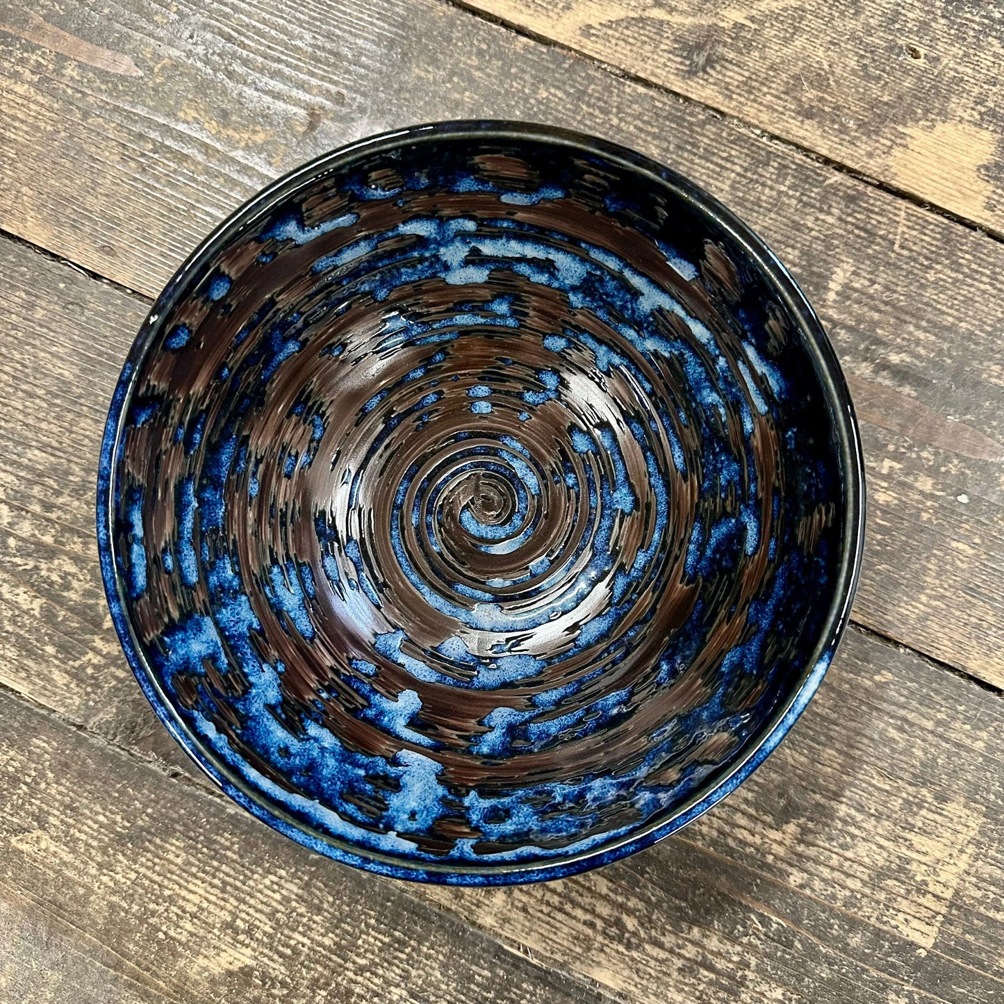 
                  
                    The Udon Bowl - Copper Swirl, a handmade Minoyaki porcelain bowl featuring a glossy copper swirl pattern, rests on a rustic wooden surface.
                  
                