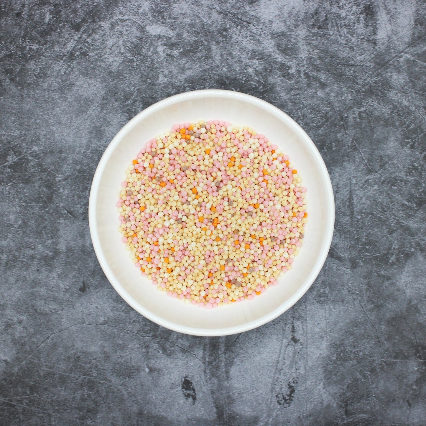 
                  
                    A white plate filled with Japanese Rice Crackers - Bubu Arare Goshiki, small and multicolored, sits on a gray textured surface.
                  
                