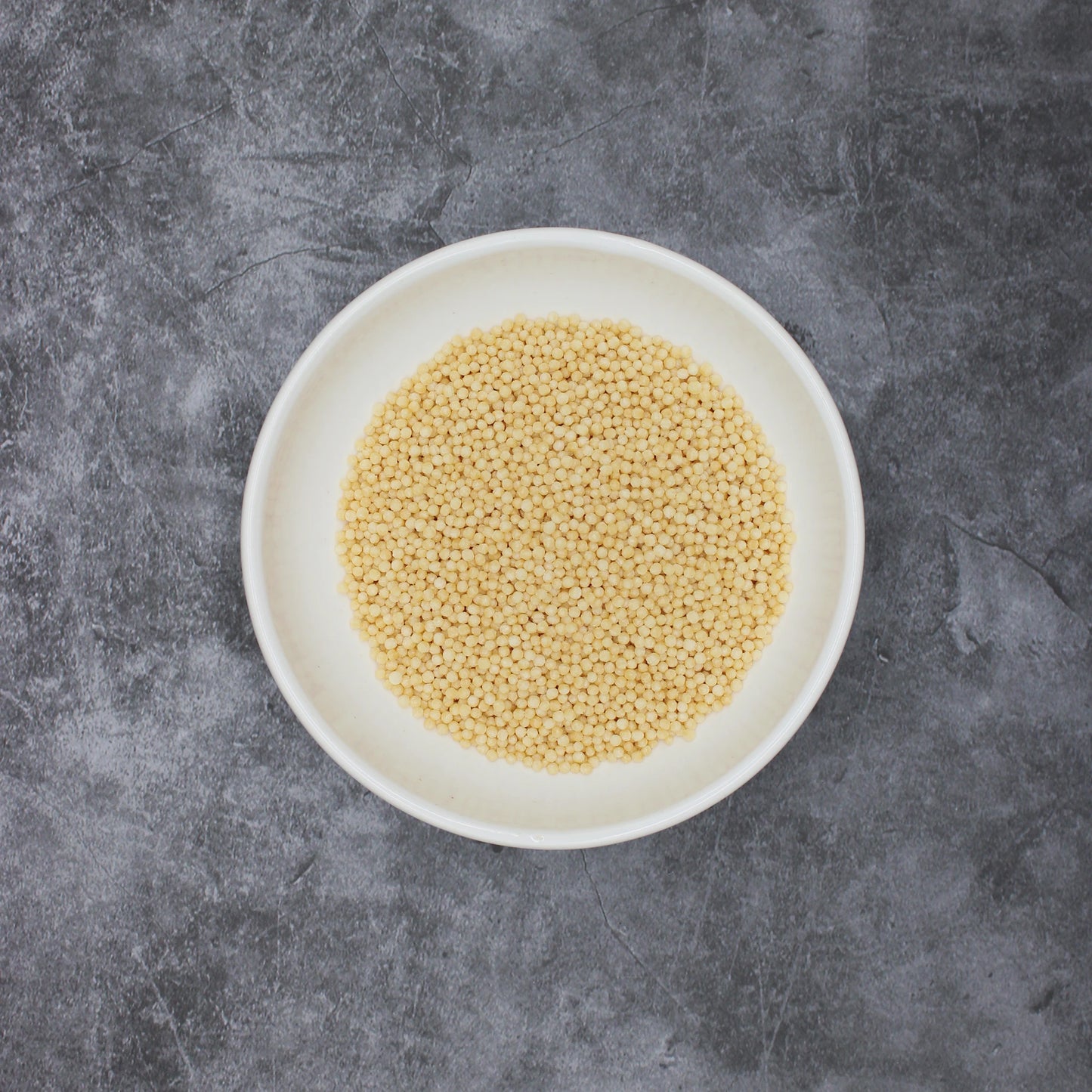 
                  
                    A white bowl of uncooked millet grains sits on a gray textured surface, evoking the delicate texture of Japanese Rice Crackers - Bubu Arare Plain.
                  
                