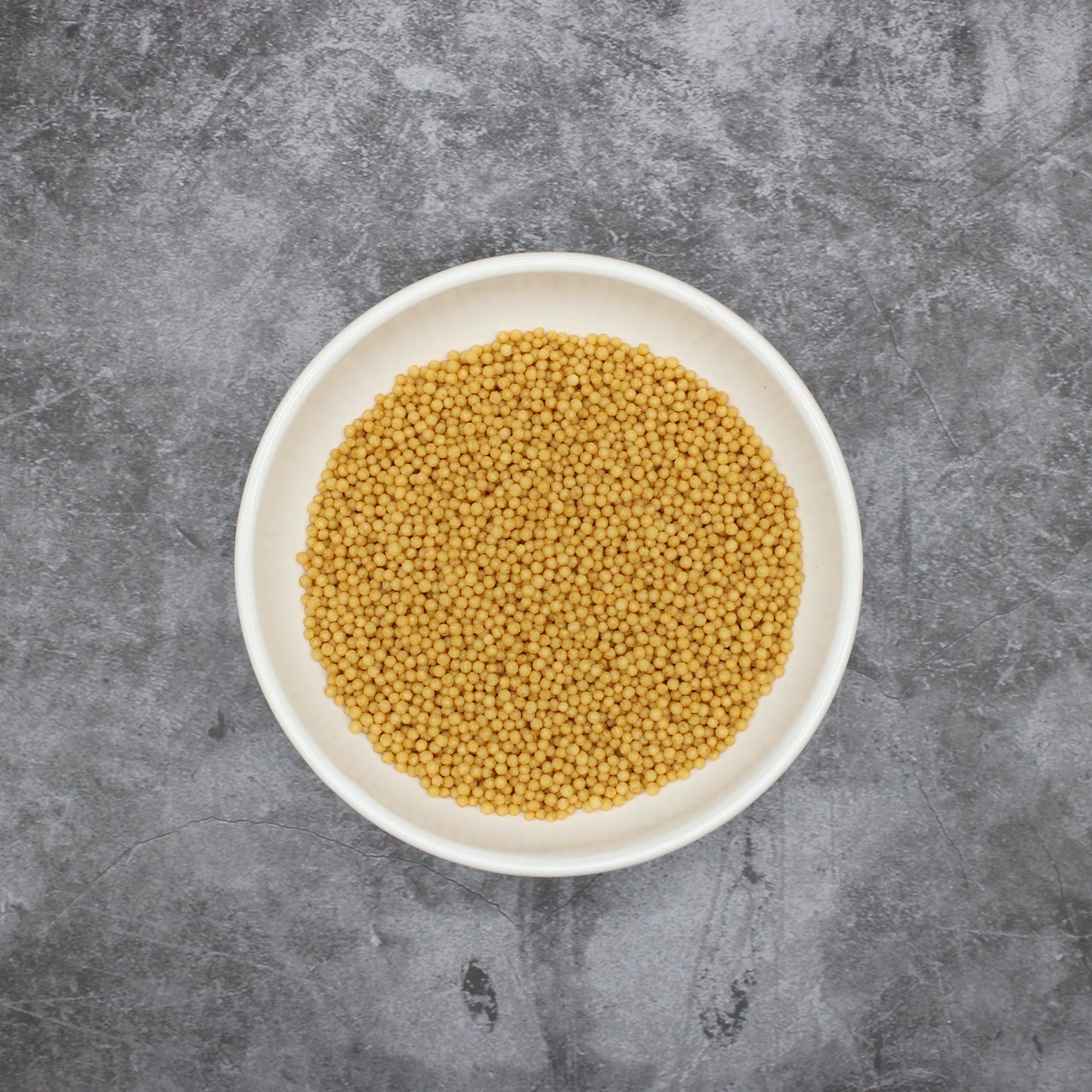 
                  
                    A white bowl filled with yellow mustard seeds sits on a gray textured surface, next to scattered Japanese Rice Crackers - Bubu Arare Wasabi.
                  
                