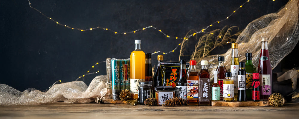 A variety of Japanese bottled and packaged food products are arranged on a wooden surface, with decorative lights and fabric in the background.