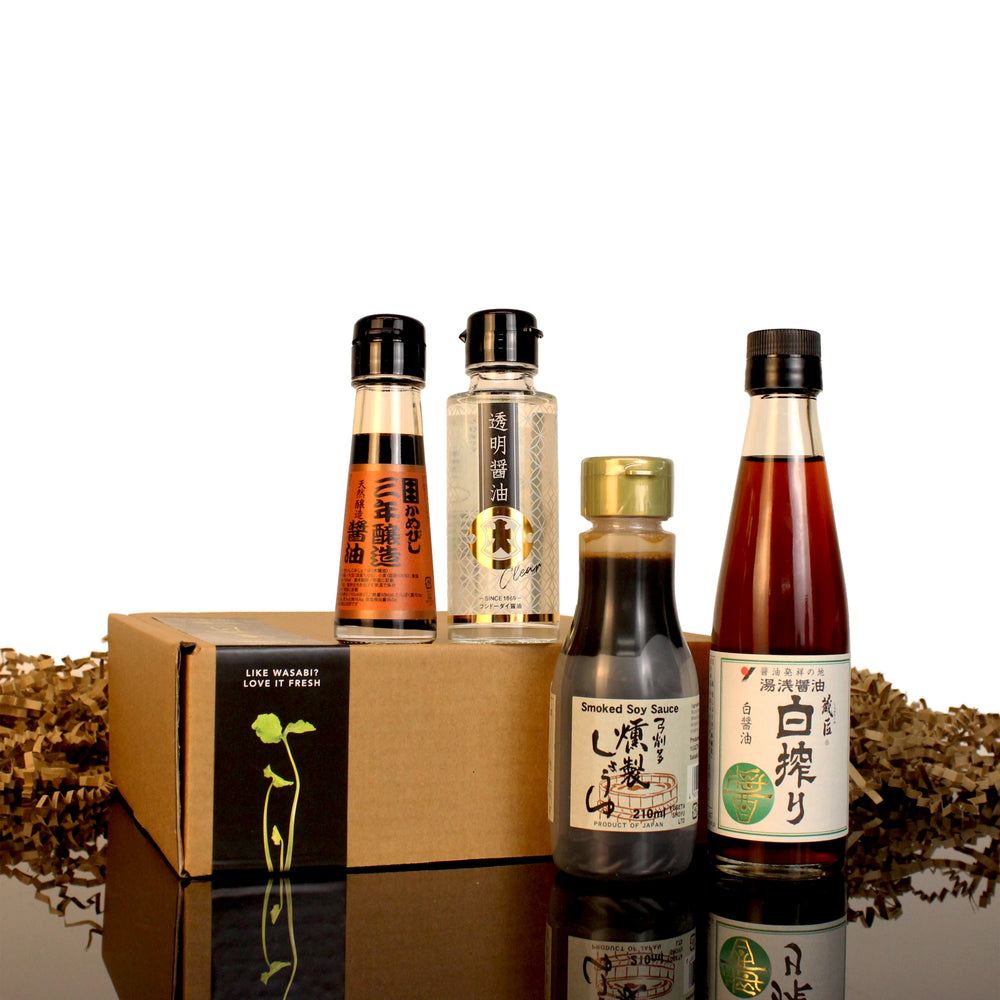 Four bottles from the Connoisseurs Soy Sauce Collection, including Japanese soy sauce and smoked soy sauce, stand upright before a brown box with a green wasabi graphic on a reflective black surface.