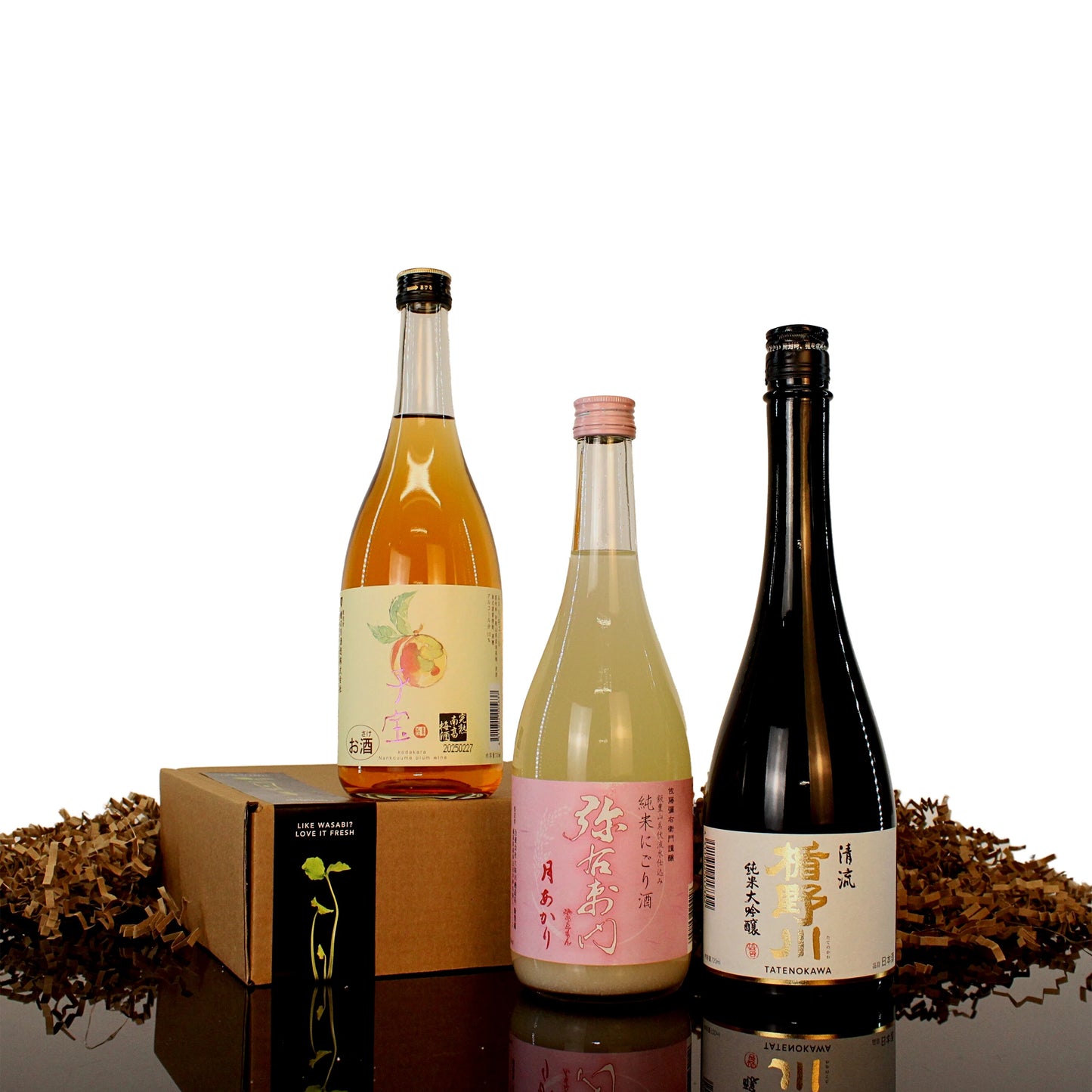 Three bottles of Japanese sake with unique labels stand upright beside the Deluxe Sake Selection gift box and shredded packing material on a black reflective surface.