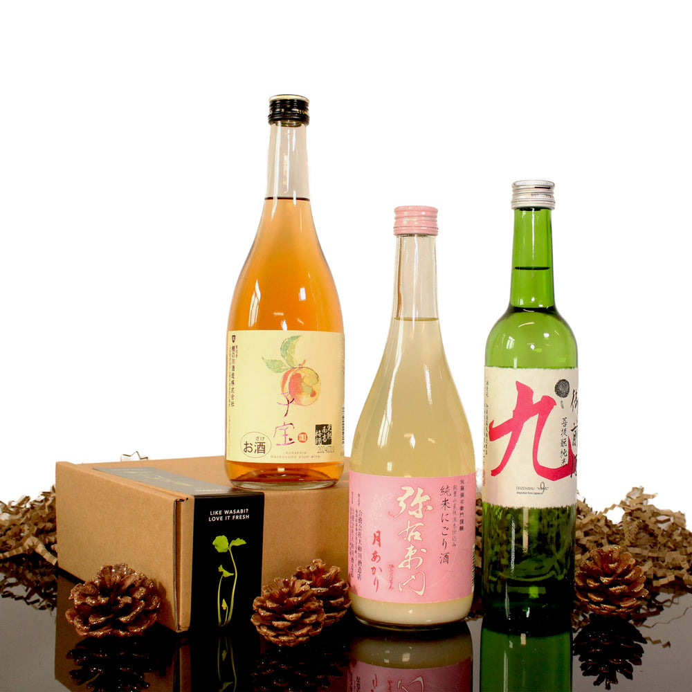 Three bottles from the Deluxe Sake Selection stand upright beside a brown box and pinecones on a reflective surface, with colorful labels visible against a white background.