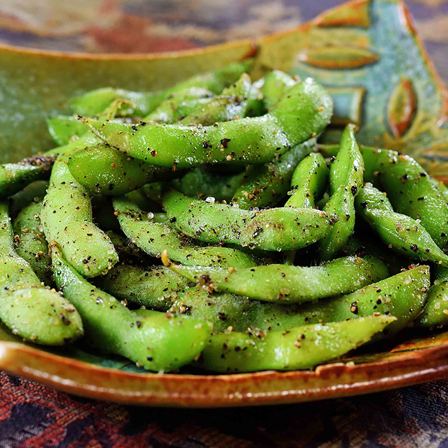 Enjoy a delicious and nutritious snack with edamame pods, harvested from Edamame Bean Plants x 3, lightly seasoned with salt and black pepper on a decorative green and brown dish.