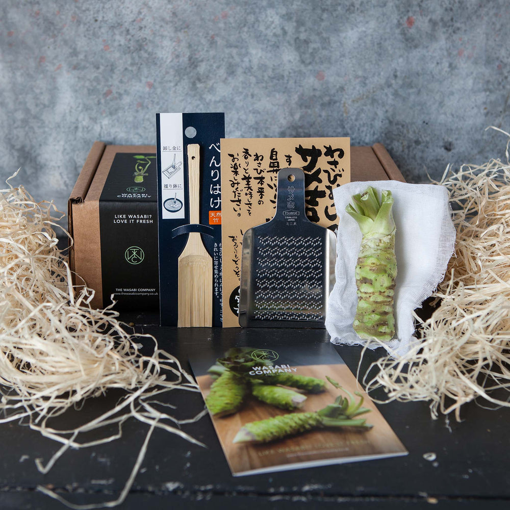 The Fresh Wasabi Starter Kit features a fresh wasabi root, metal grater, bamboo brush, and instruction card in stylish packaging with straw filling, all showcased on a black surface.