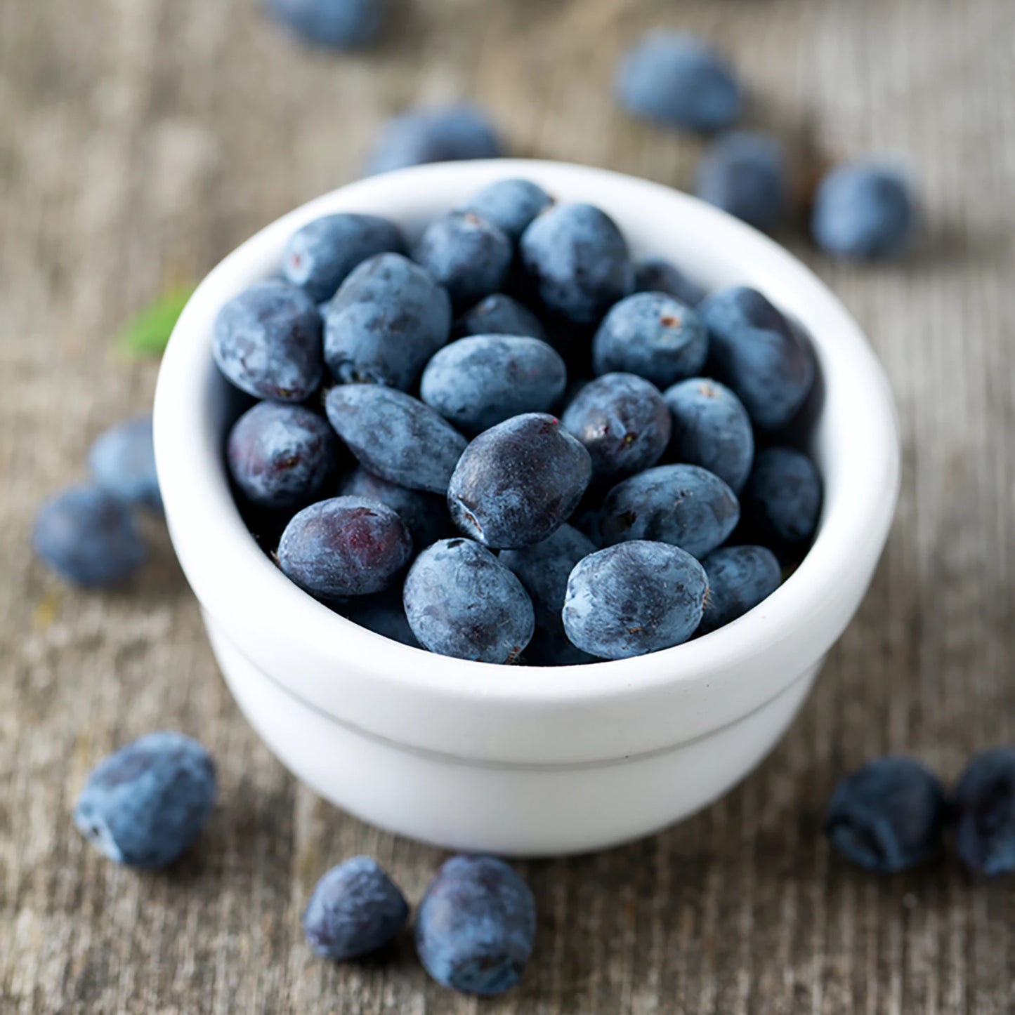 A white bowl with fresh, nutrient-rich Honeyberry Plant - Giants Heart berries sits on a wooden surface, with a few antioxidant-rich berries scattered around.