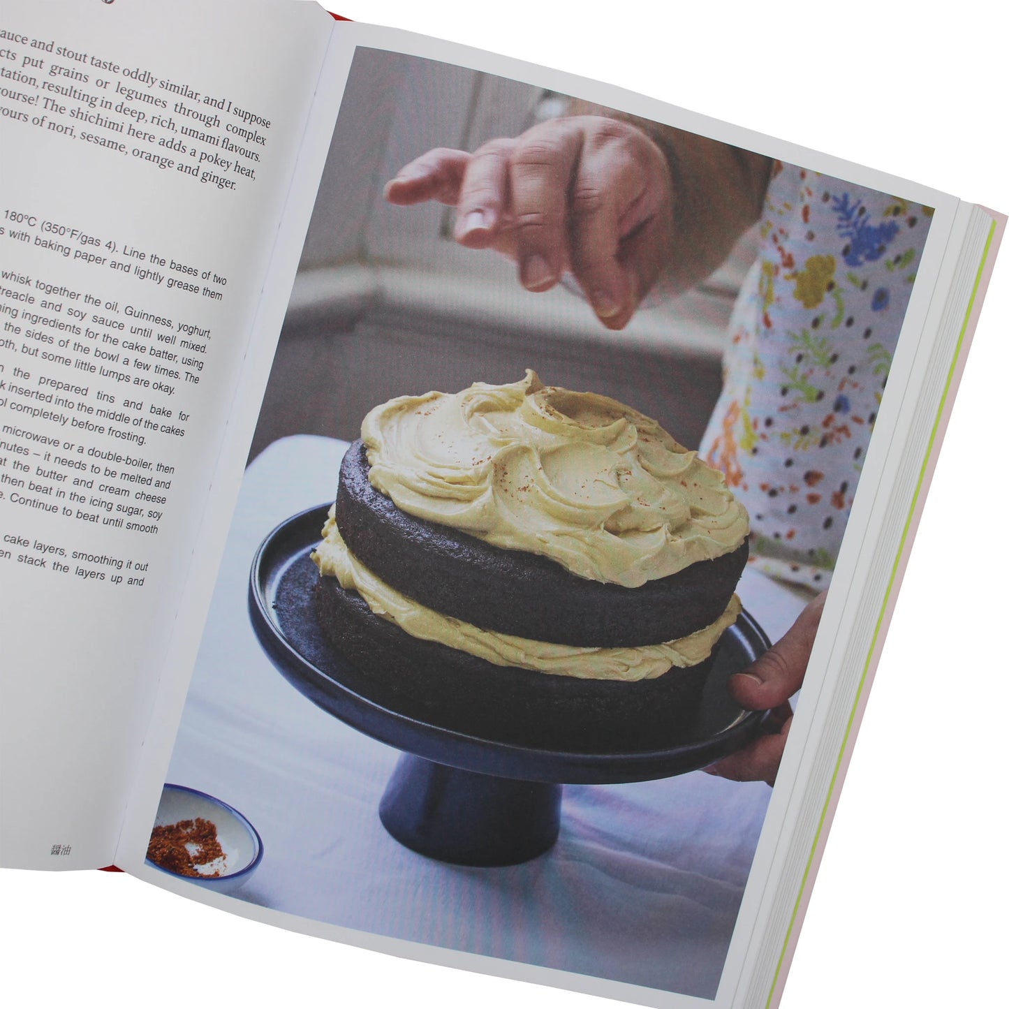 
                  
                    A hand sprinkles seasoning over a two-layer chocolate cake on a dark stand, featured in the Japaneasy Kitchen by Tim Anderson cookbook.
                  
                