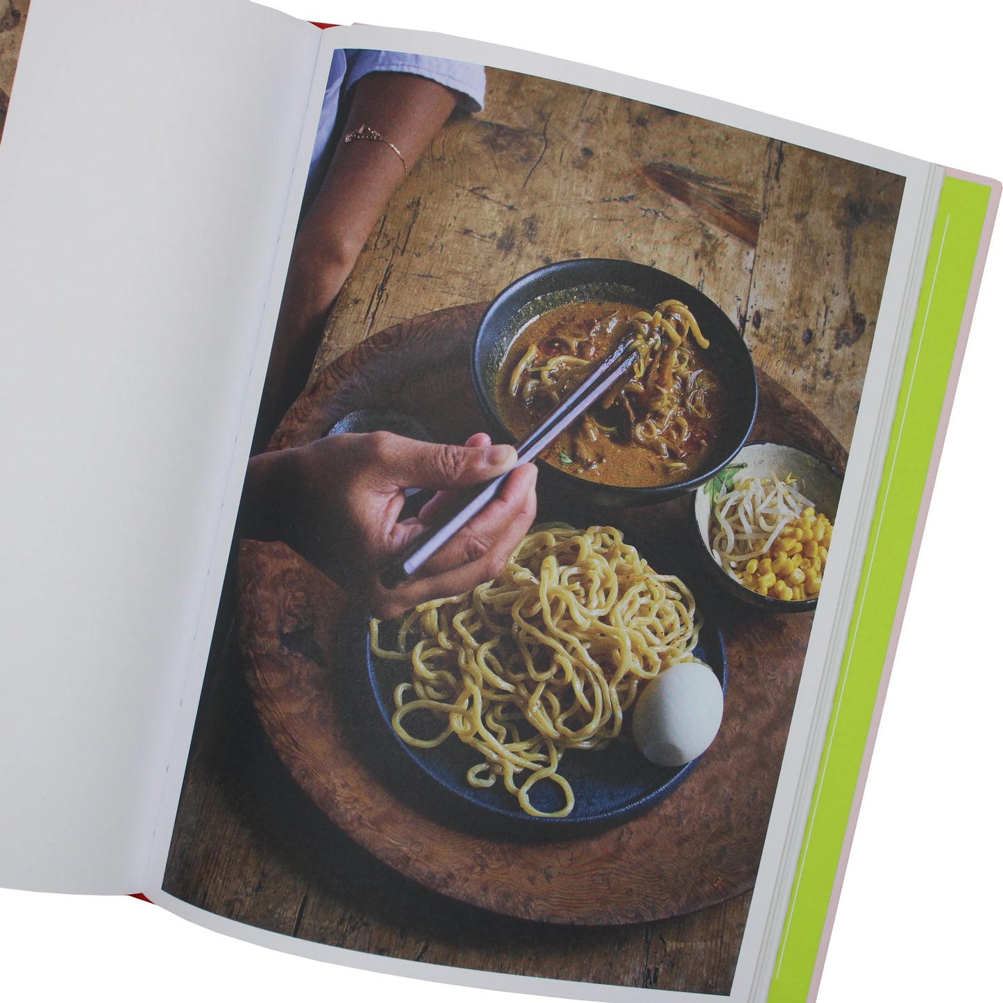 
                  
                    A person uses chopsticks to eat Japanese pantry noodles from a Japaneasy Kitchen by Tim Anderson bowl, with a side plate of noodles, a hard-boiled egg, corn, and bean sprouts on a wooden table.
                  
                