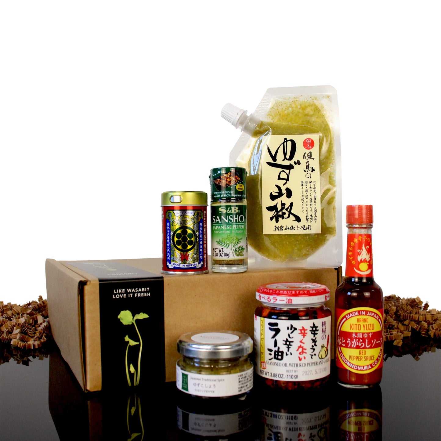 A Japanese Spice Set consisting of a pouch of yuzu kosho, bottles of chili oil and red pepper sauce, and small jars of Sansho pepper, arranged in front of a cardboard shipping box.