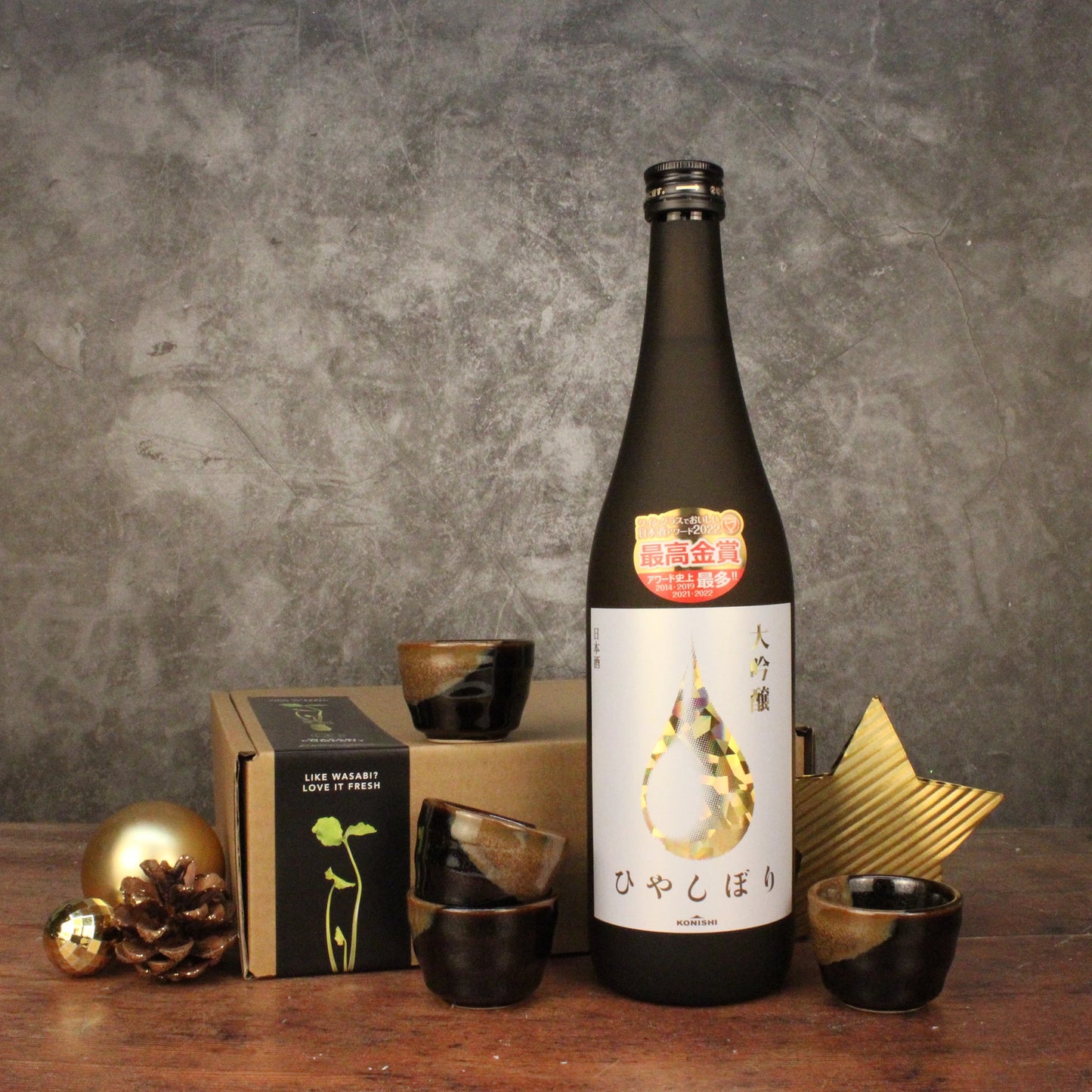 A bottle of Konishi Gold Sake with Japanese text is displayed on a table alongside its set of four sake cups, the product box, and gold decorative ornaments.