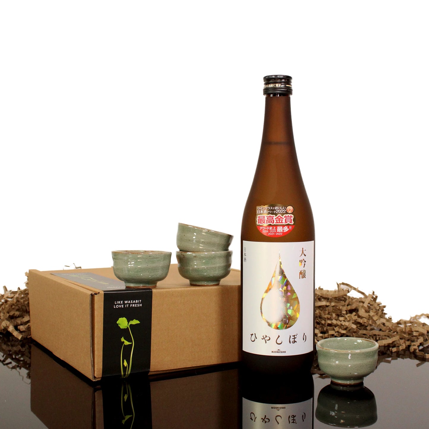 A bottle of Konishi Gold Sake with Japanese labeling is displayed next to a set of four Japanese ceramic sake cups and a cardboard box with shredded packing material on a reflective black surface.