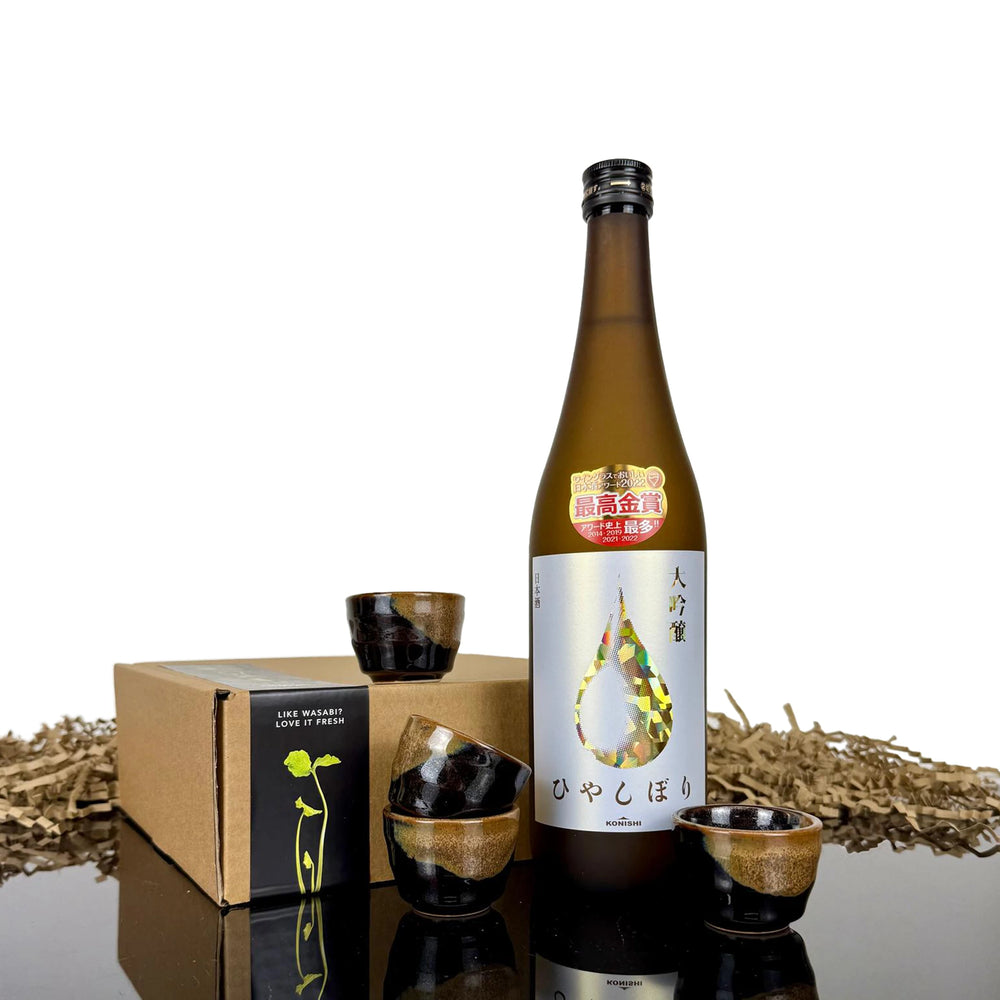 Konishi Gold Sake, paired with four Japanese ceramic sake cups, is displayed beside a brown box against a white background with packaging material behind.