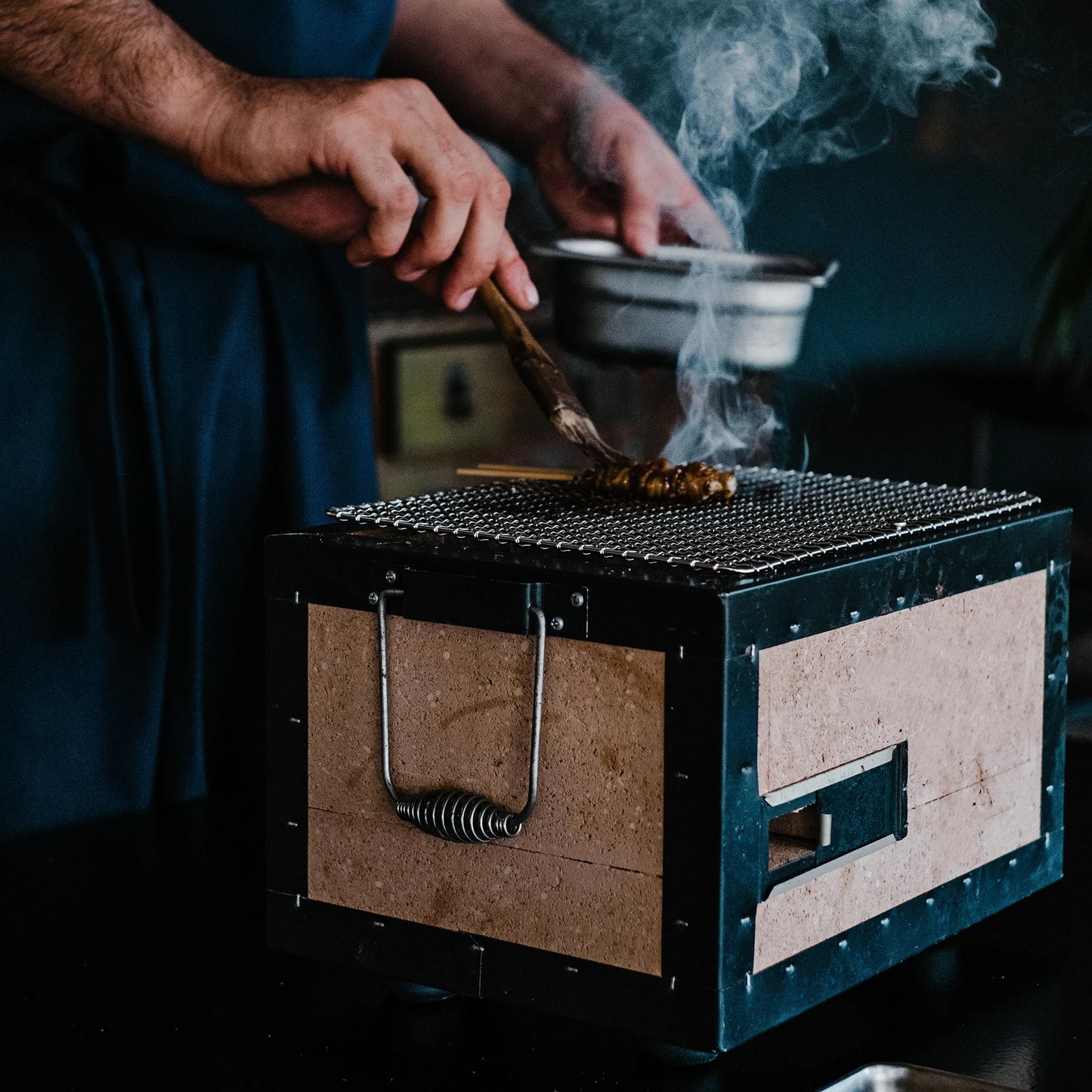 
                  
                    Someone grills skewers on the 31cm Konro Grill Table Barbecue, smoke rising as they brush sauce or oil onto the food.
                  
                