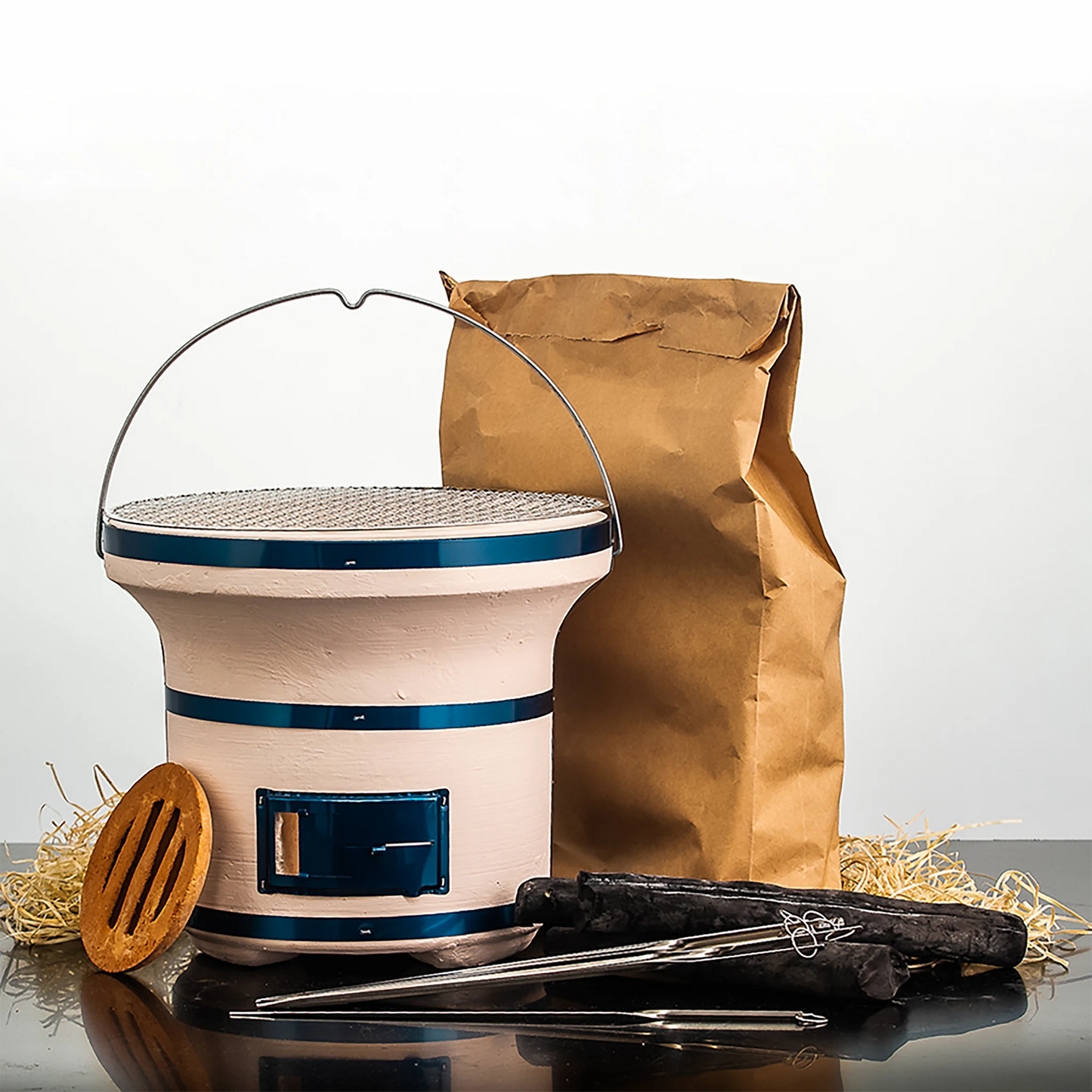 The Shichirin Table Barbecue Set includes a portable clay charcoal stove, metal grate, wooden spatula, professional steel skewers, Binchotan charcoal pieces, and a brown paper bag—all arranged on a reflective surface.