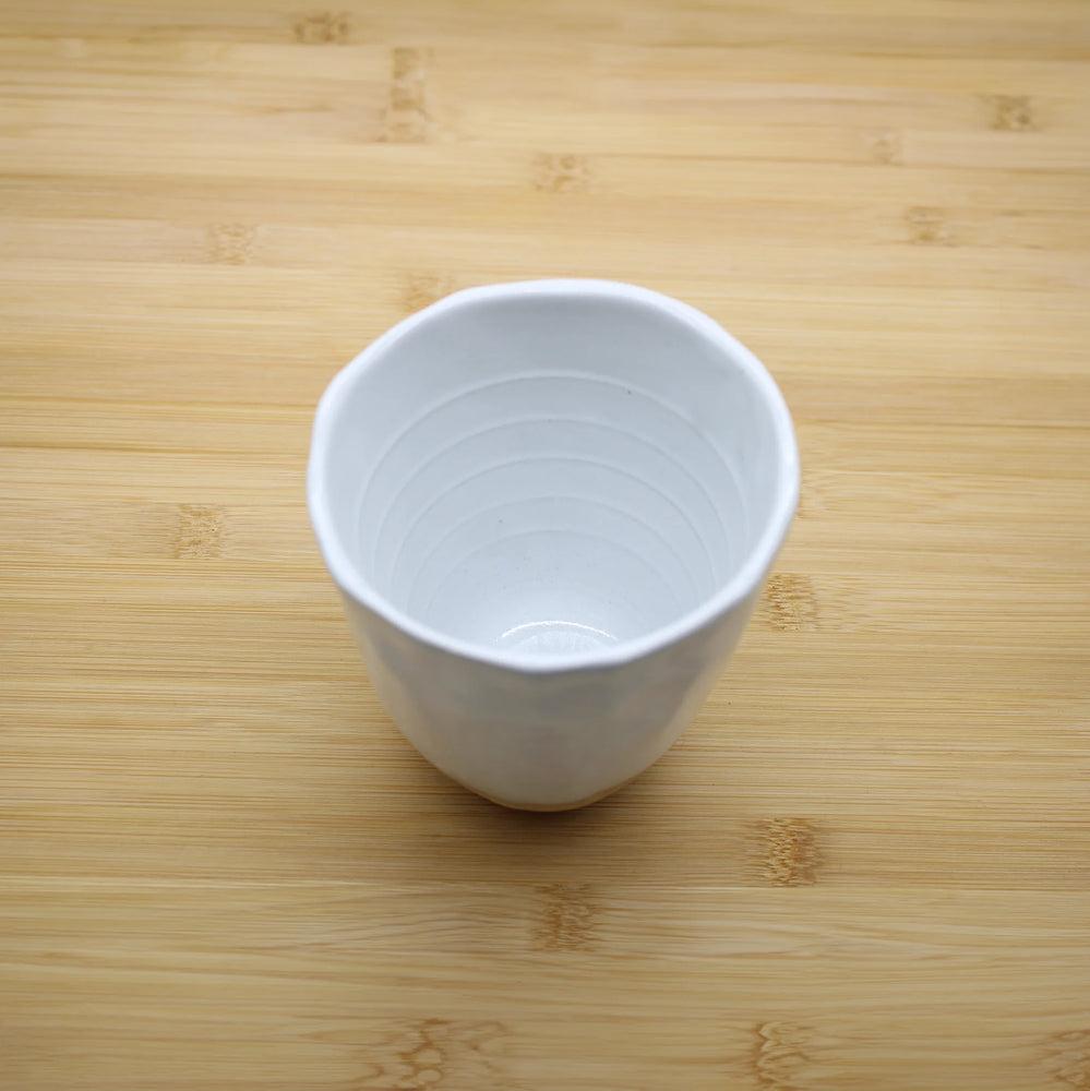 
                  
                    The Lopsided Mug - White And Bisque, featuring a slightly irregular rim and earthenware base, sits on a light wooden surface and reflects Japanese craftsmanship.
                  
                