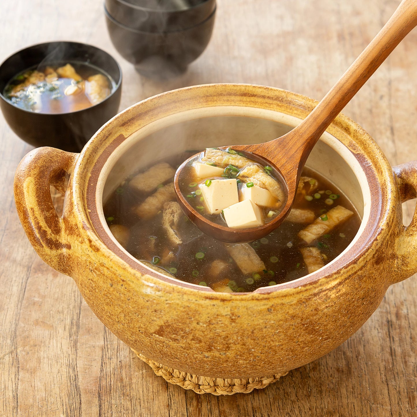 
                  
                    A Miso Soup Pot filled with tofu, seaweed, and green onions is served with a wooden ladle and two smaller bowls of soup in the background on a wooden table.
                  
                