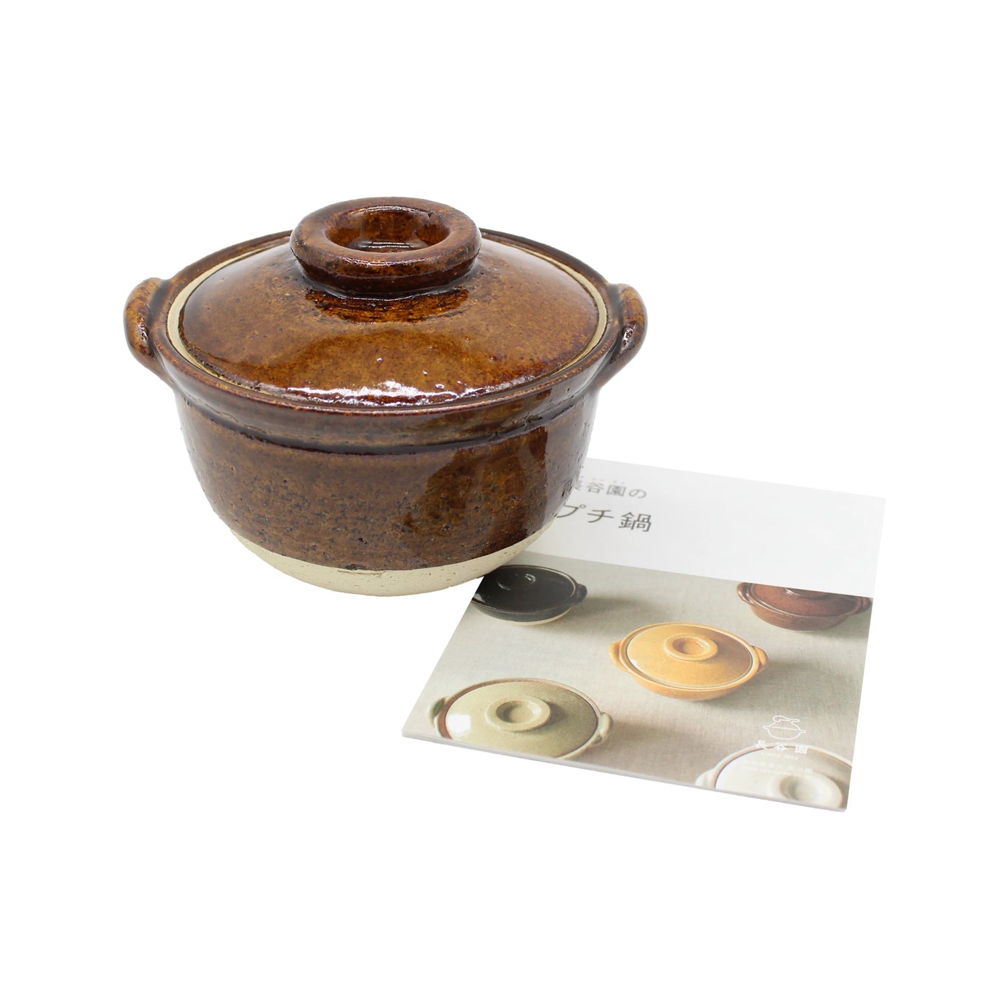 
                  
                    A Mini Donabe Pot - Ame, a brown ceramic Iga ware pot with a lid and handles, is placed next to a booklet that features images of similar Japanese handmade tableware on its cover.
                  
                