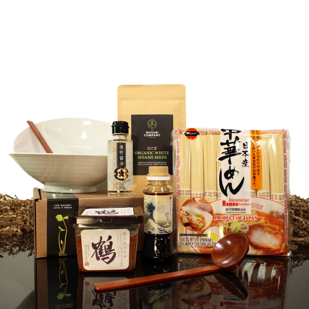 A ramen kit including Japanese noodles, seasoning packs, a large bowl, a spoon, jars of sauces, sesame seeds, and a bottle of sauce, all arranged on a reflective surface.
