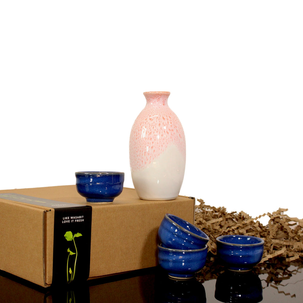 The Sake Carafe with Cups set features a white and pink carafe with four blue cups, displayed on and beside a cardboard box against brown packing paper.