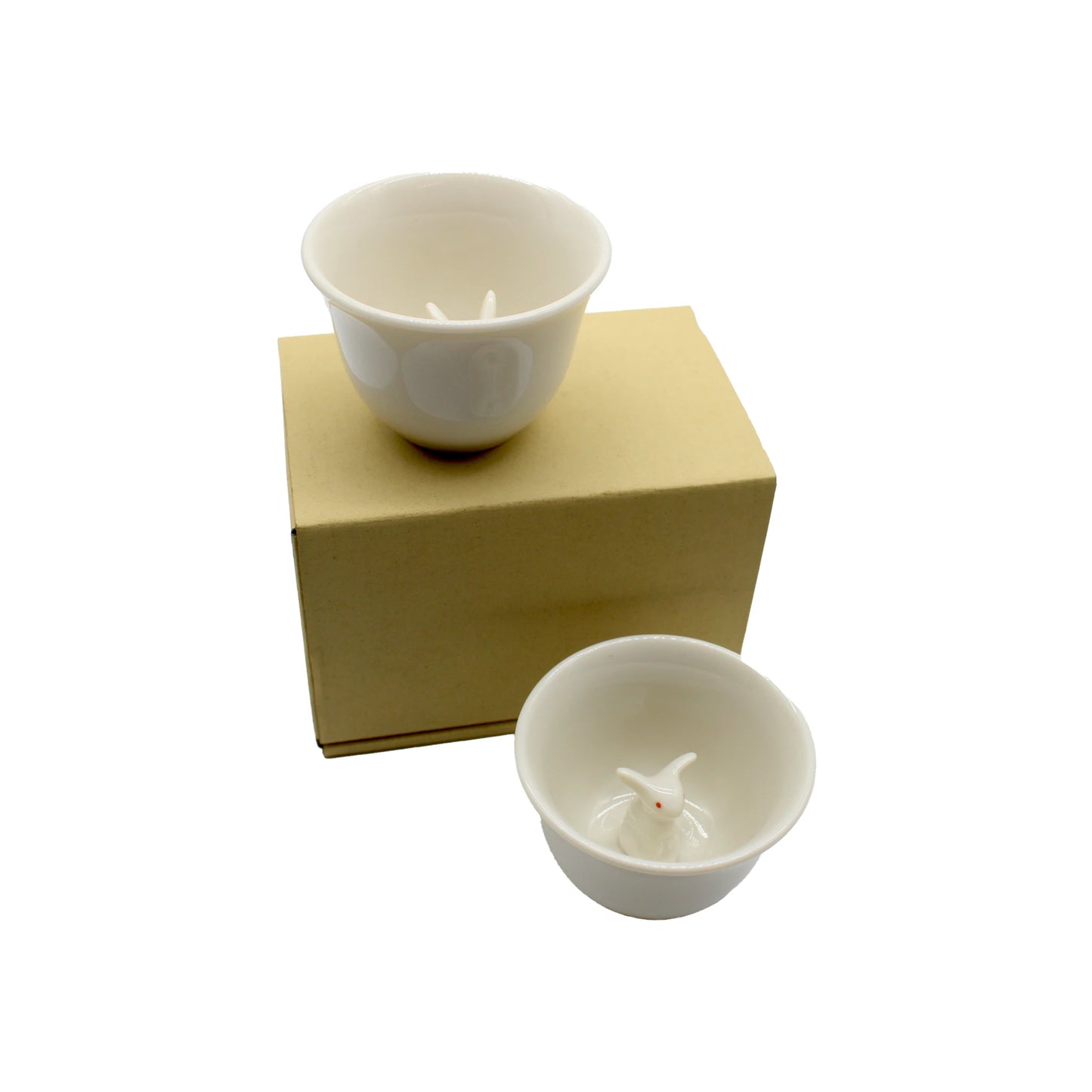 
                  
                    The "Sake Cup Boxed Set - Rabbit Design" features two white ceramic cups inspired by Japanese ceramics, each adorned with a small bunny figurine inside, and is shown displayed on and beside its plain cardboard box against a white background.
                  
                
