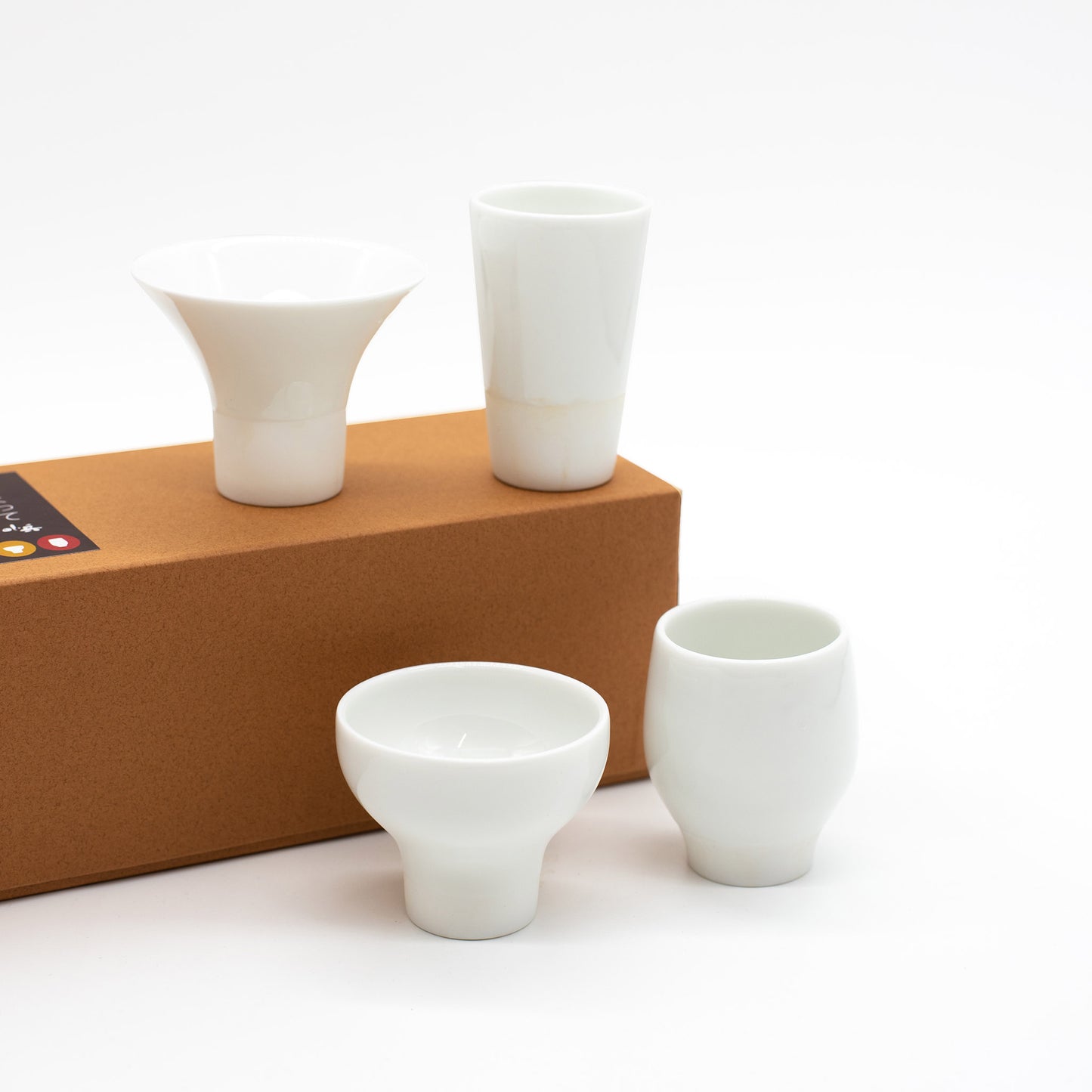 The Sake Cups Tasting Boxed Set features four uniquely shaped white porcelain cups, highlighting Kaneko Kohyo craftsmanship, displayed on and beside a rectangular brown box against a white background.