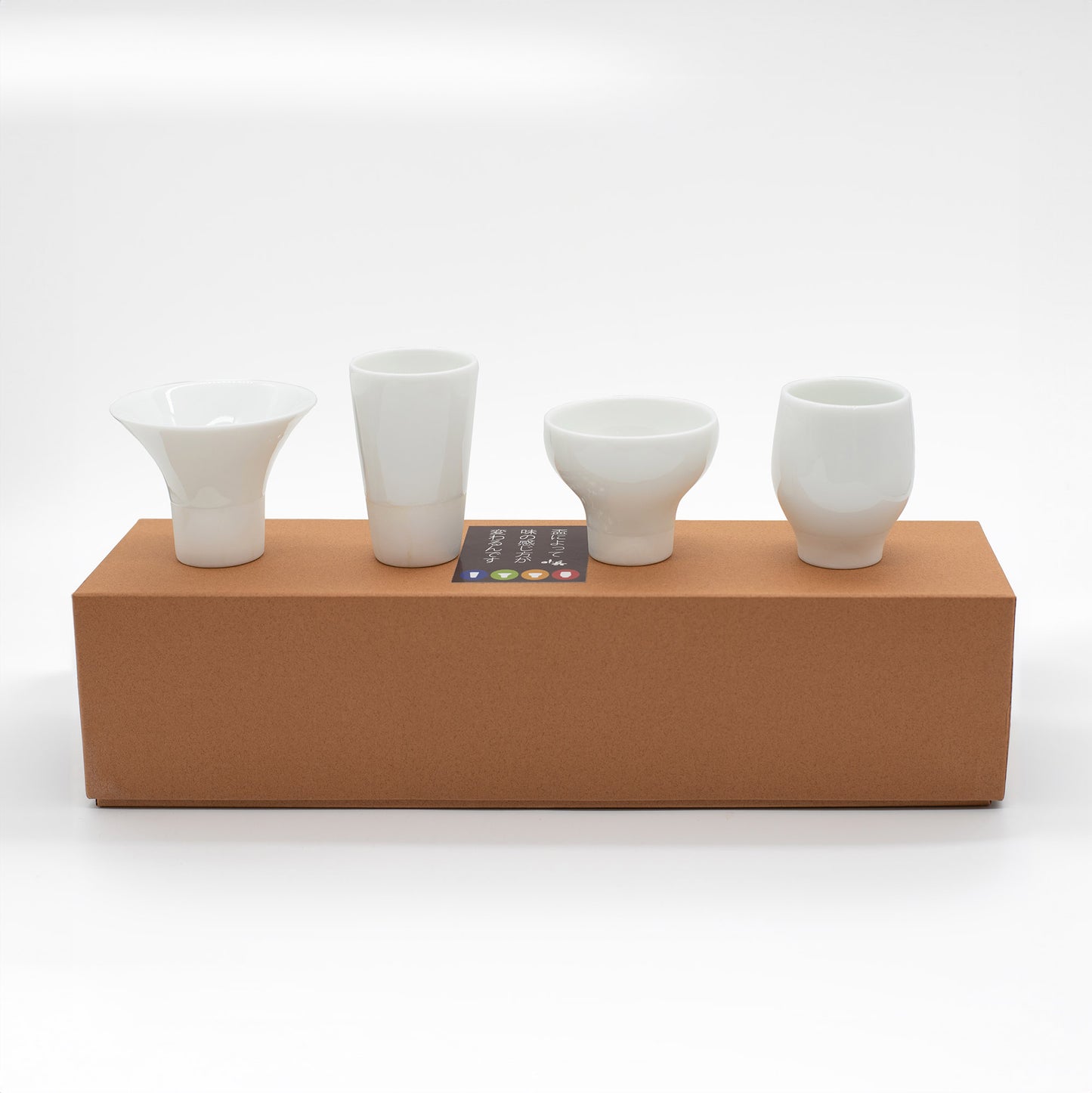 The Sake Cups Tasting Boxed Set features four unique white ceramic cups, beautifully crafted and displayed in a row on a brown box—ideal for an elegant sake tasting experience.