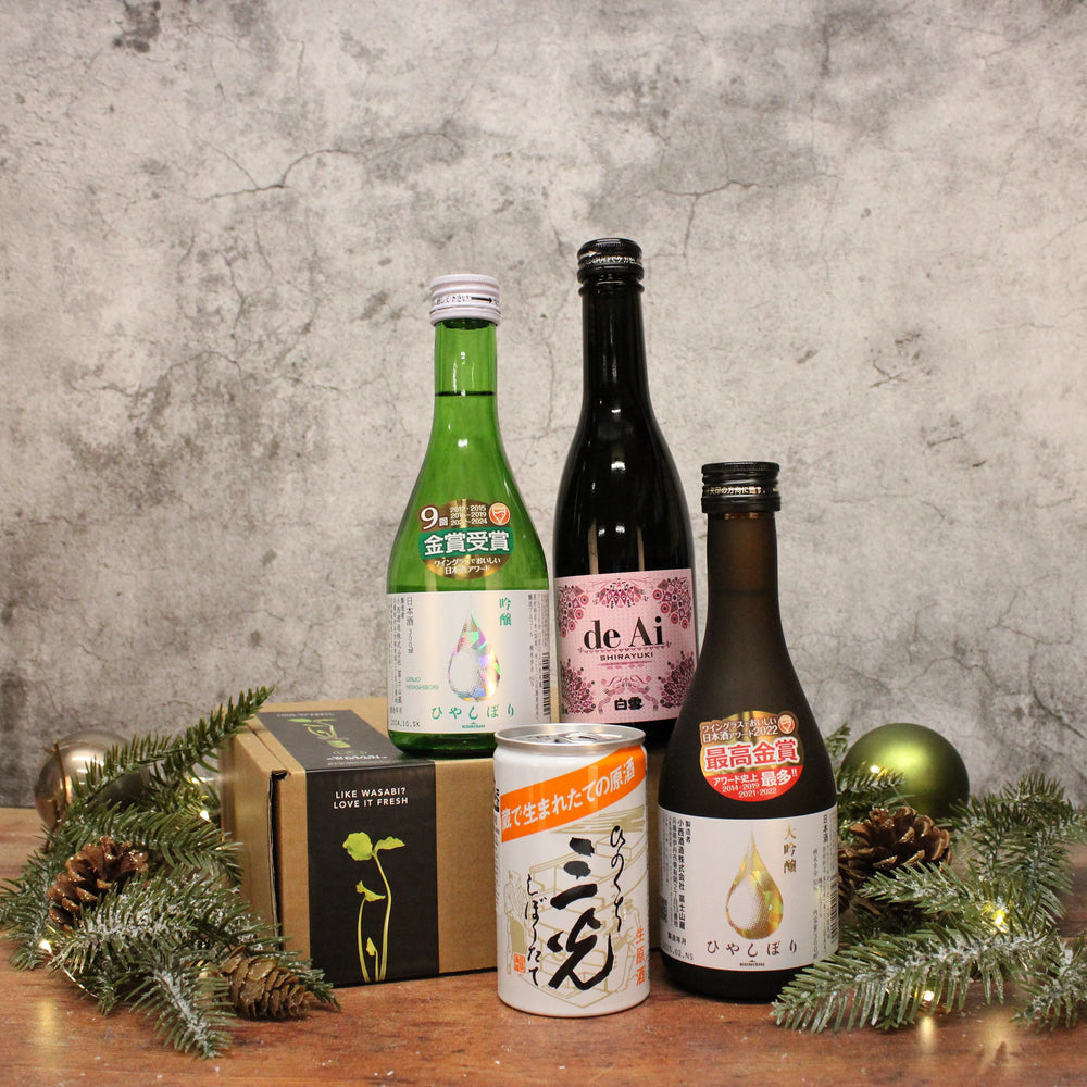 Sake Selection: Four premium sake bottles and cans, including sparkling sake, beautifully arranged with a green gift box, pine branches, and ornaments on a textured background.