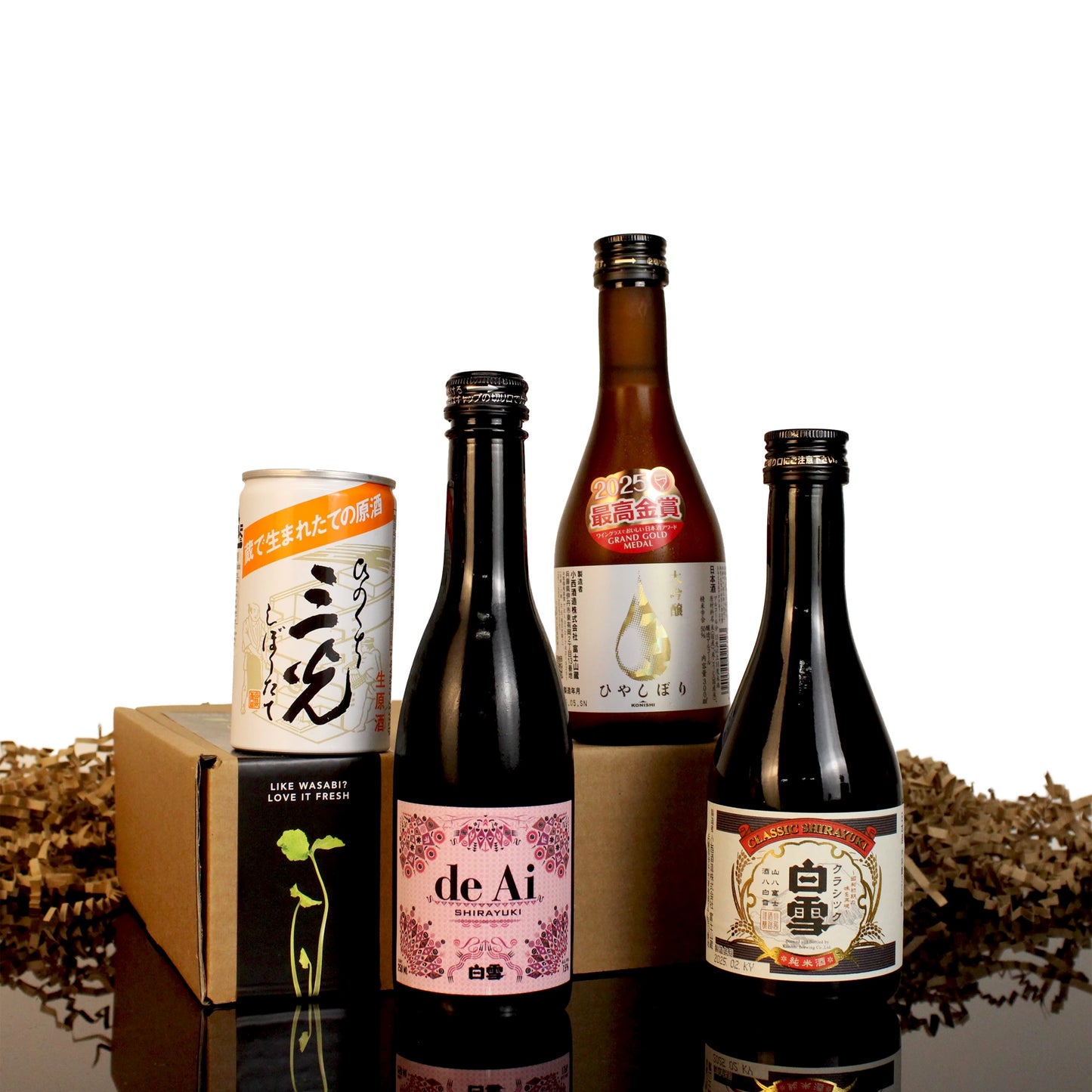 The Sake Selection features five premium Japanese sake bottles and cans, artfully arranged with crinkle paper atop brown cardboard boxes, all set on a reflective surface.
