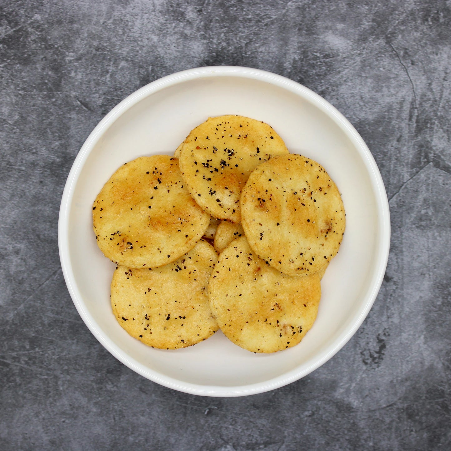 
                  
                    A white bowl filled with Japanese Rice Crackers - Black Pepper Senbei 90g sits on a gray textured surface, showcasing round, golden-brown rice crackers seasoned with black pepper for a tasty twist on classic snacks.
                  
                