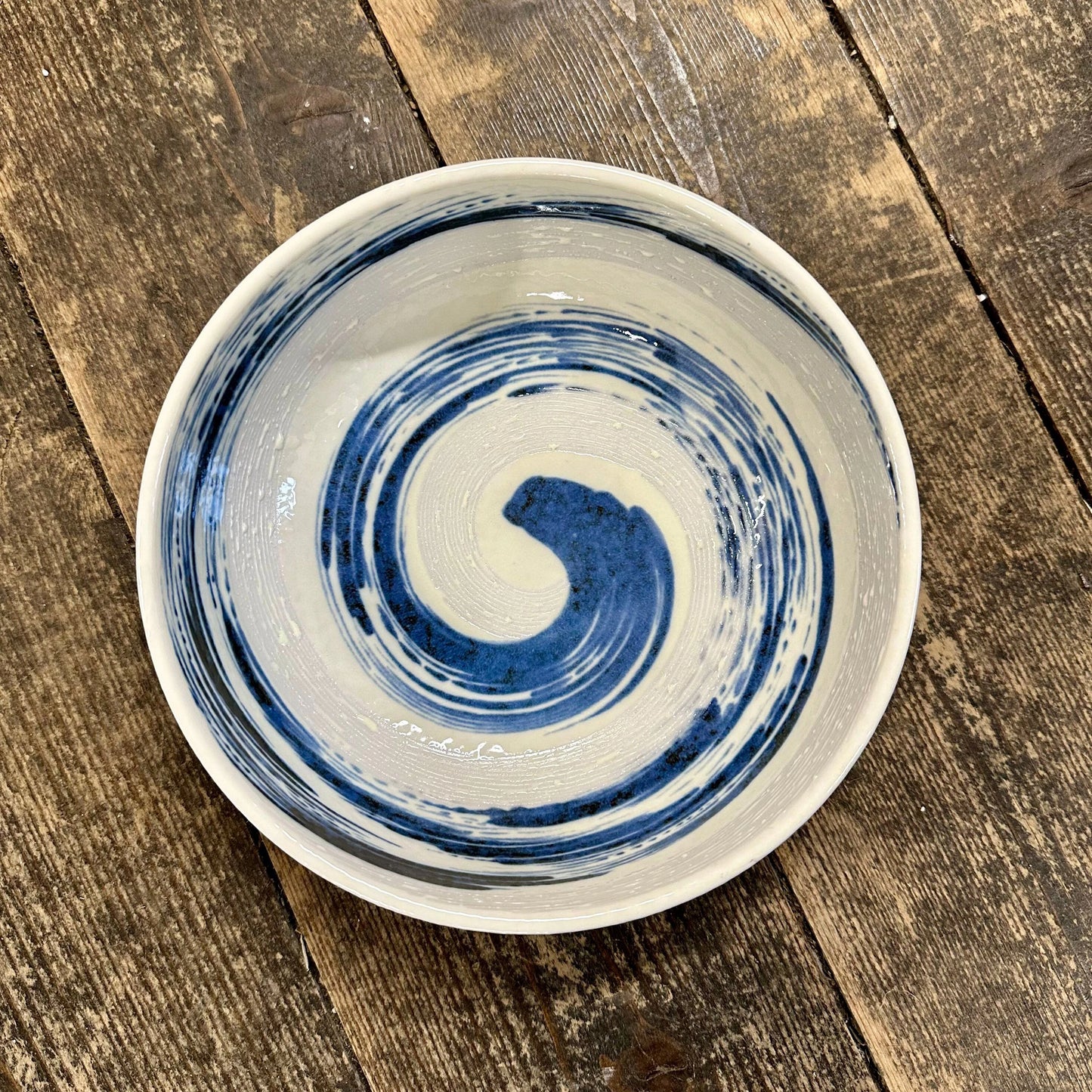 
                  
                    The Open Bowl - Blue Swirl, crafted from Minoyaki porcelain with a blue spiral design on white, sits on a rustic wooden surface—an elegant showcase of Japanese pottery.
                  
                