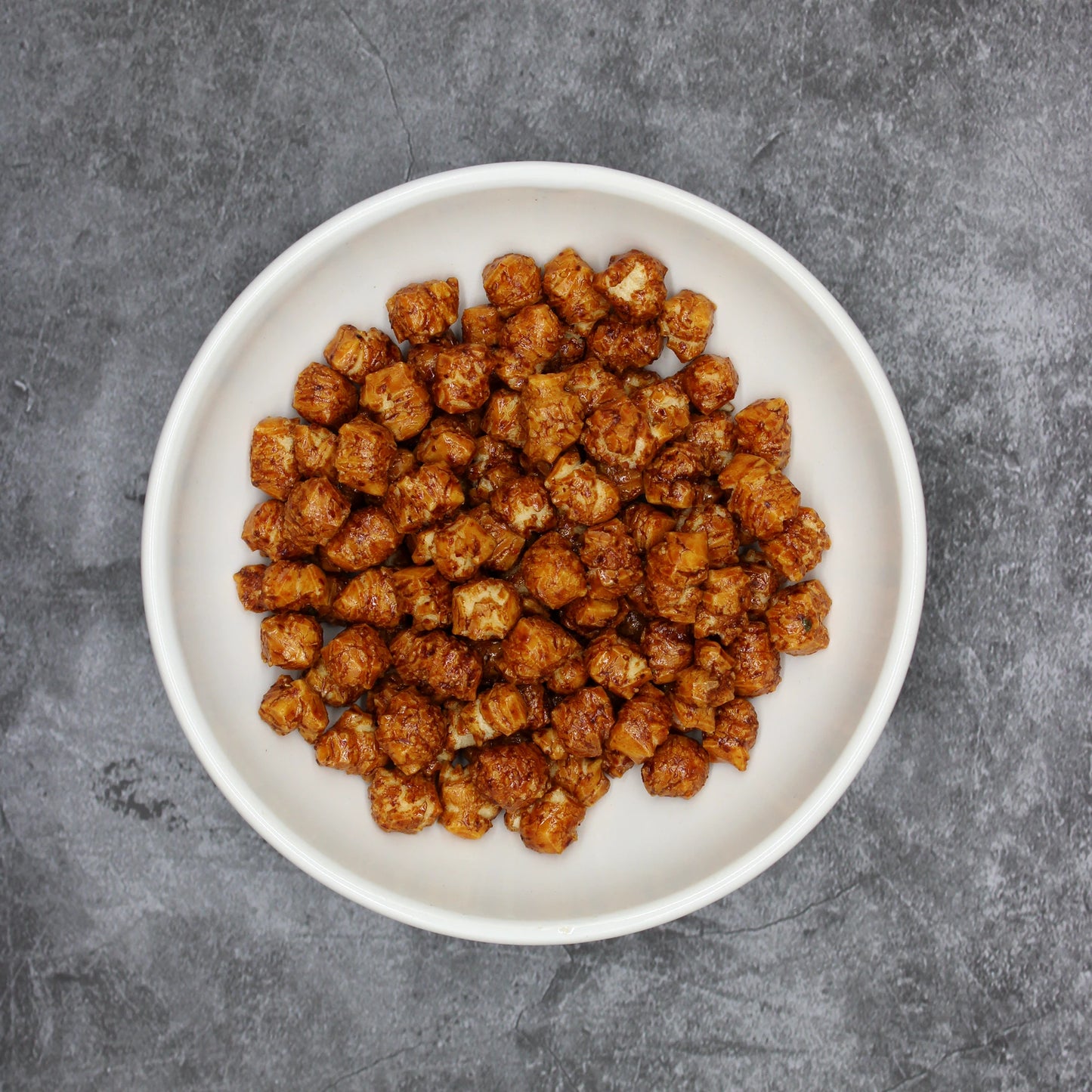 
                  
                    A white bowl of Japanese Rice Crackers - Shichimi Kozo 80g, featuring small, brown, cubed crackers dusted with shichimi togarashi, rests on a gray textured surface for a savory snack.
                  
                