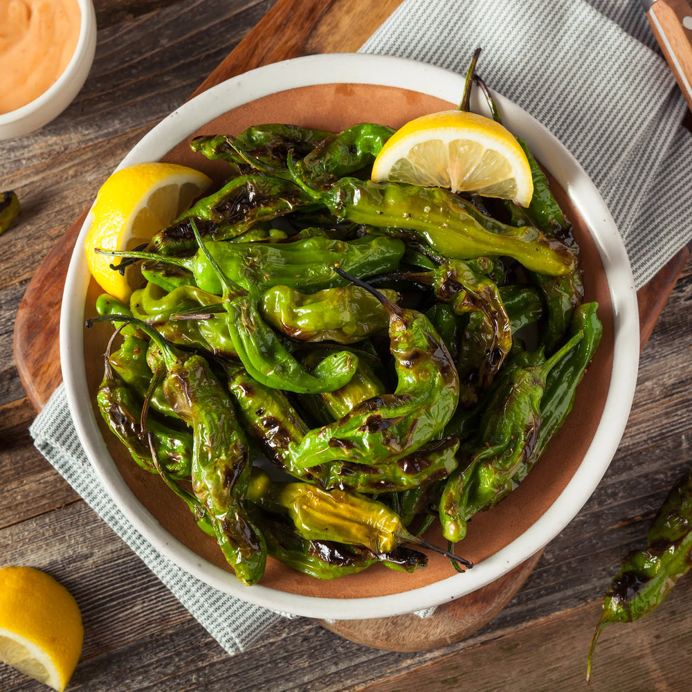 
                  
                    A harvest from Shishito Pepper Plants x 3—mild, sweet peppers, charred and served with lemon wedges and dipping sauce—rests on a wooden table near a striped cloth.
                  
                
