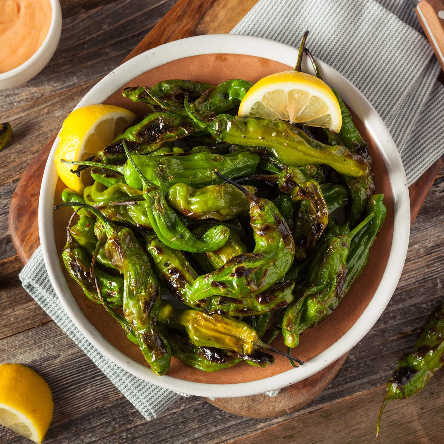 
                  
                    A harvest from Shishito Pepper Plants x 3—mild, sweet peppers, charred and served with lemon wedges and dipping sauce—rests on a wooden table near a striped cloth.
                  
                