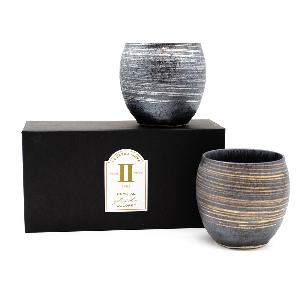 A boxed set of two Sho-Chu Cups, crafted in Minoyaki porcelain, features exquisite Japanese craftsmanship. One cup rests on the black box labeled “1 Pair Cup” and “Crystal Gold & Silica Colored,” with the other placed in front.