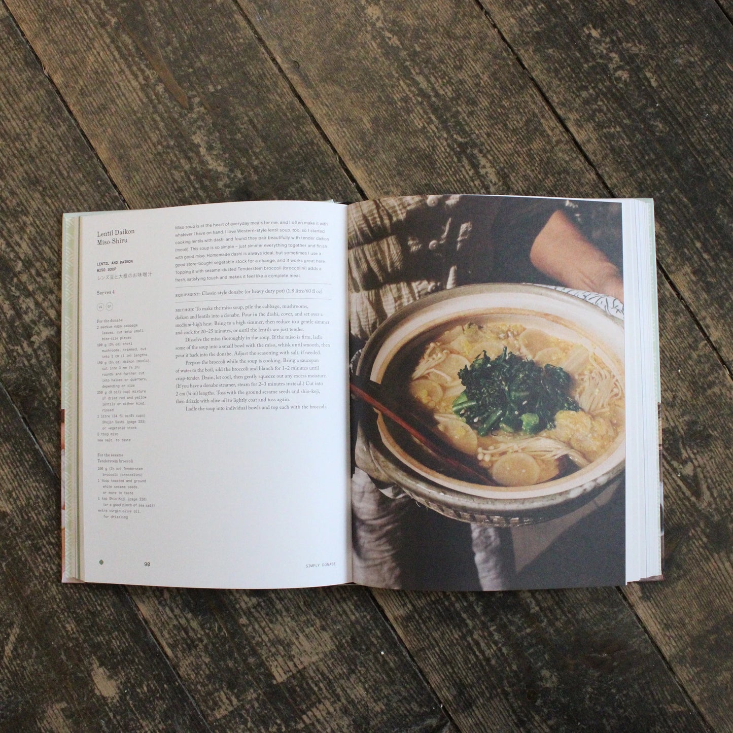
                  
                    An open copy of Simply Donabe - Japanese One Pot Recipes by Naoko Takei Moore rests on a wooden surface, showing a lentil dashi recipe on the left and a photo of noodle soup with greens in earthenware pots on the right.
                  
                
