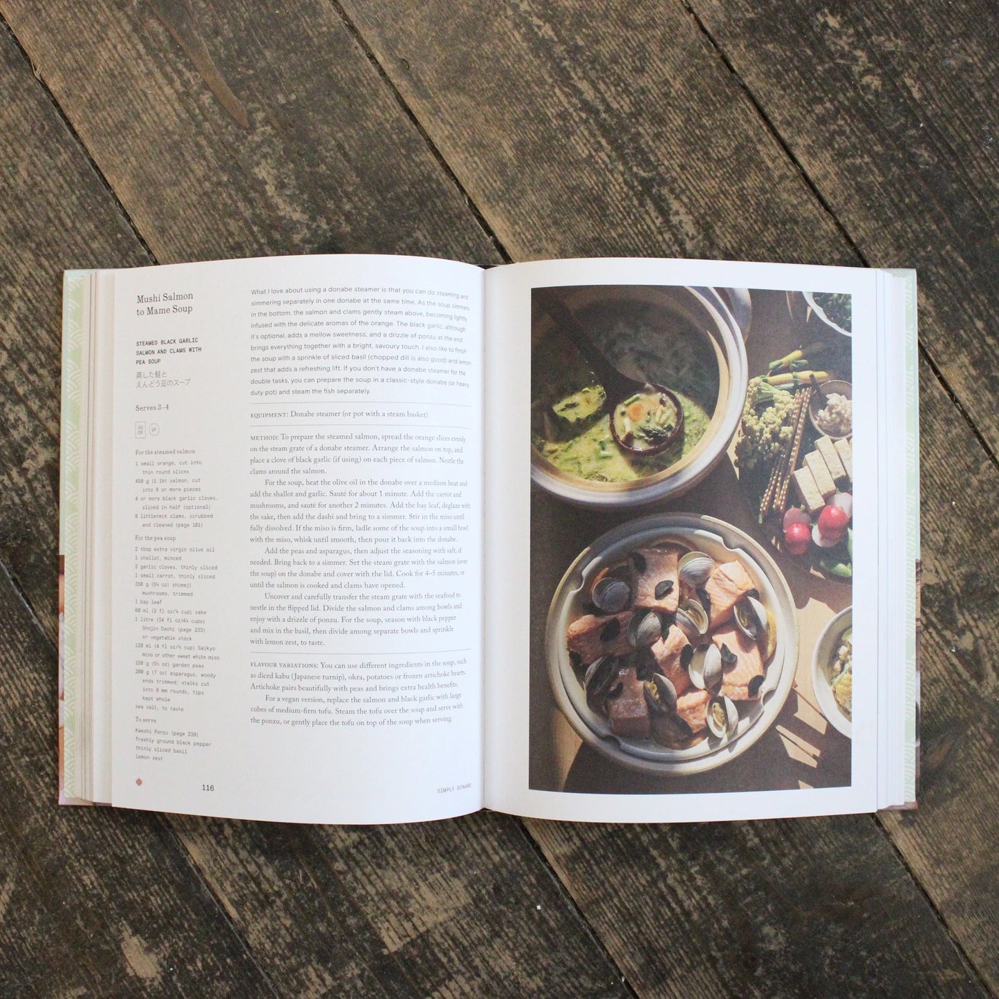 
                  
                    An open copy of "Simply Donabe - Japanese One Pot Recipes by Naoko Takei Moore" rests on a wooden floor, showing a recipe on the left page and a photo of seafood soup with vegetables in donabe pots on the right.
                  
                