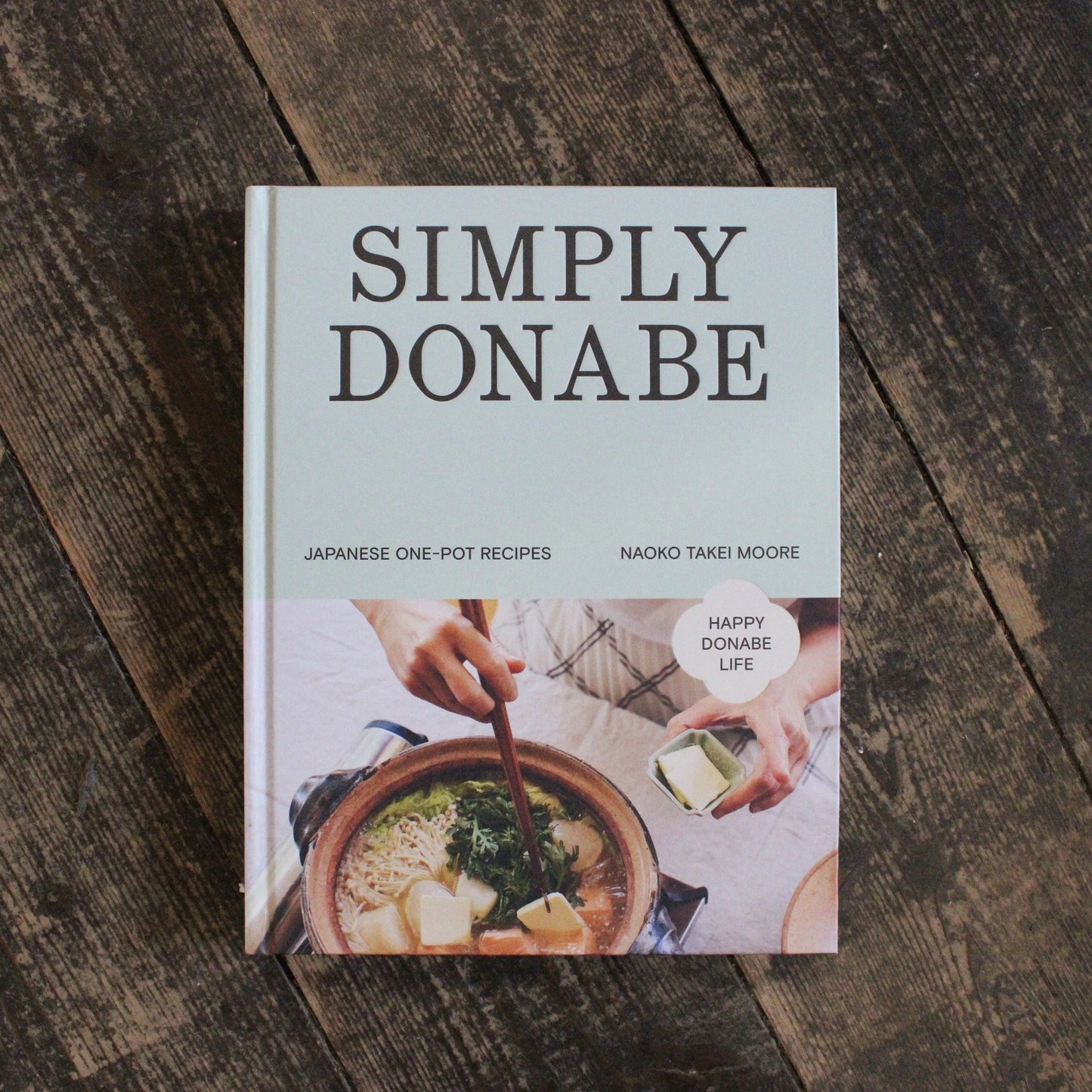 
                  
                    The cookbook "Simply Donabe - Japanese One Pot Recipes by Naoko Takei Moore" is displayed on a wooden surface.
                  
                
