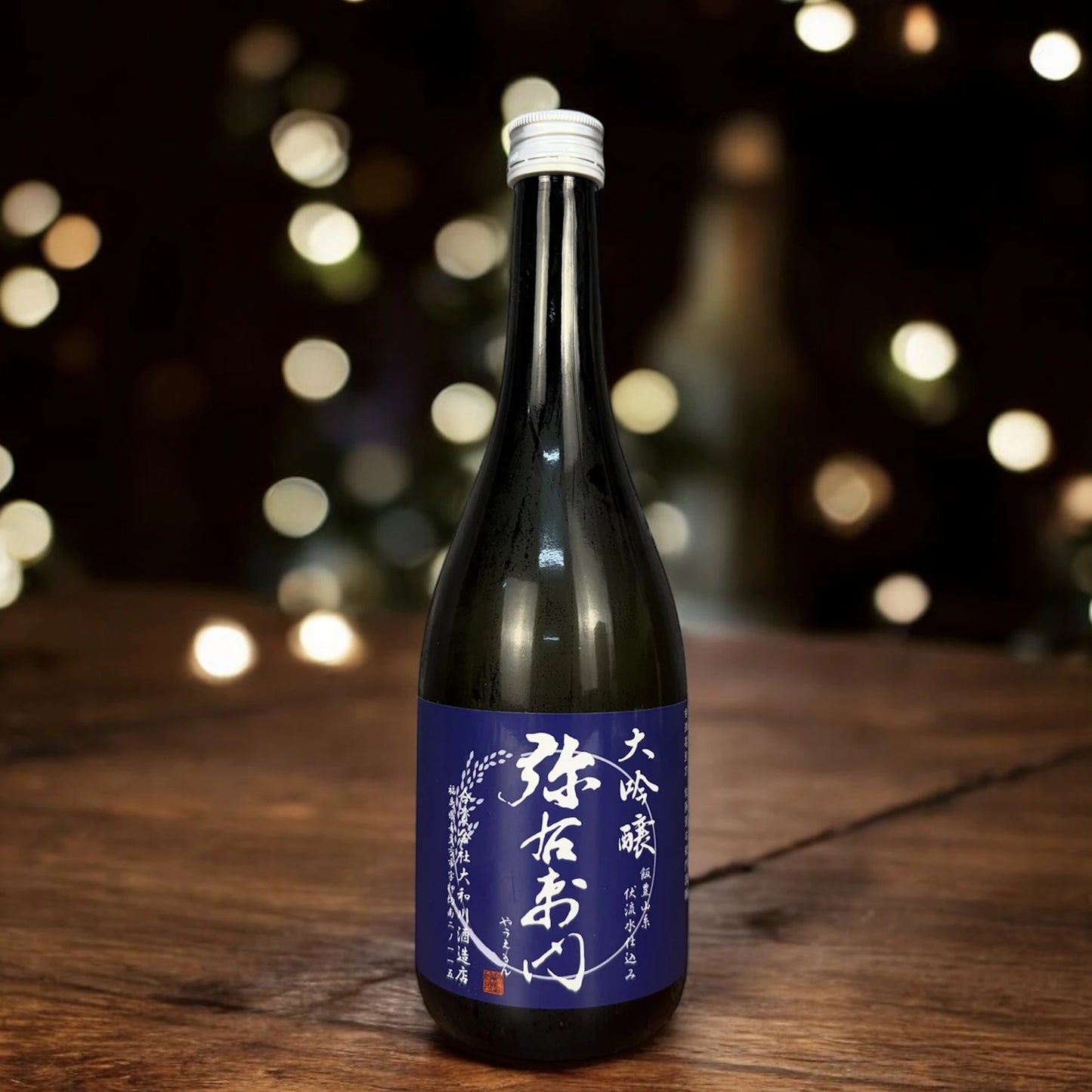 A 720ml dark glass bottle of Snow Blossom Daiginjo Sake with a blue label and white kanji sits on a wooden surface, blurred lights in the background.